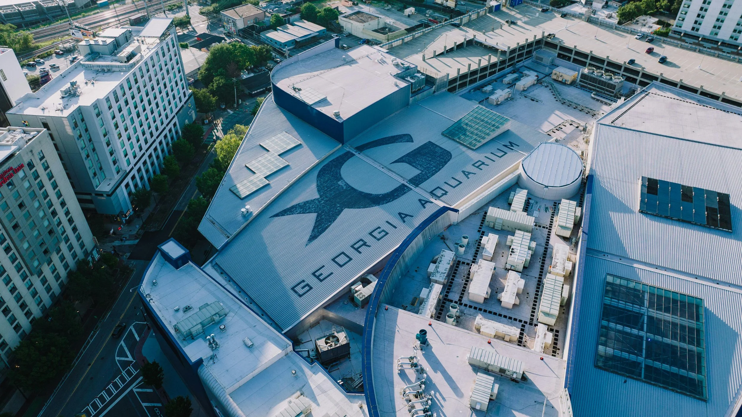 Aerial view of a city rooftop with the words 'GEORGIA AQUARIUM' and a dolphin logo painted on it, surrounded by other buildings, parking lot, and trees.