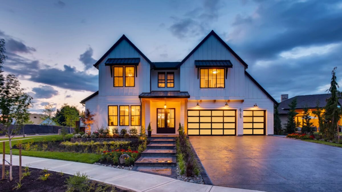 Modern two-story house with white exterior and black accents, illuminated windows, and a spacious driveway at dusk.