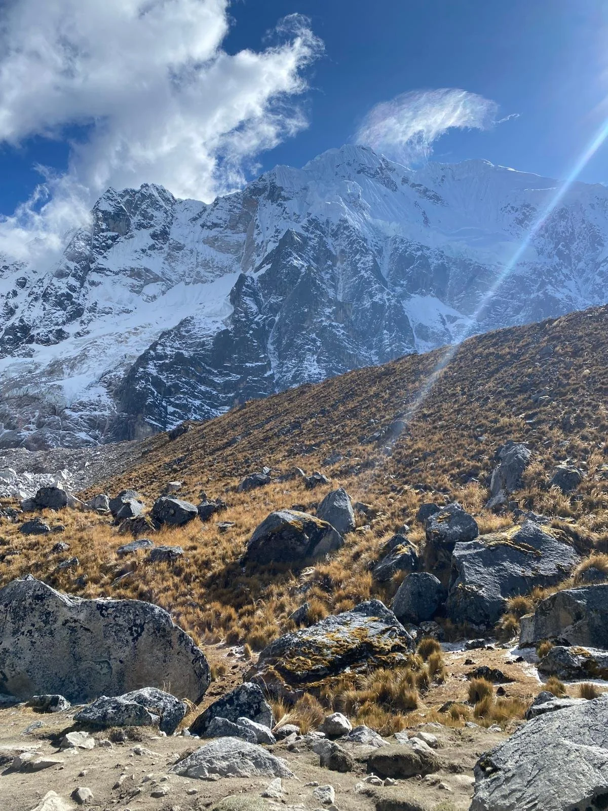 Snow-covered mountain peaks under a bright blue sky with scattered clouds, rocky and grassy hillside in the foreground with large boulders.