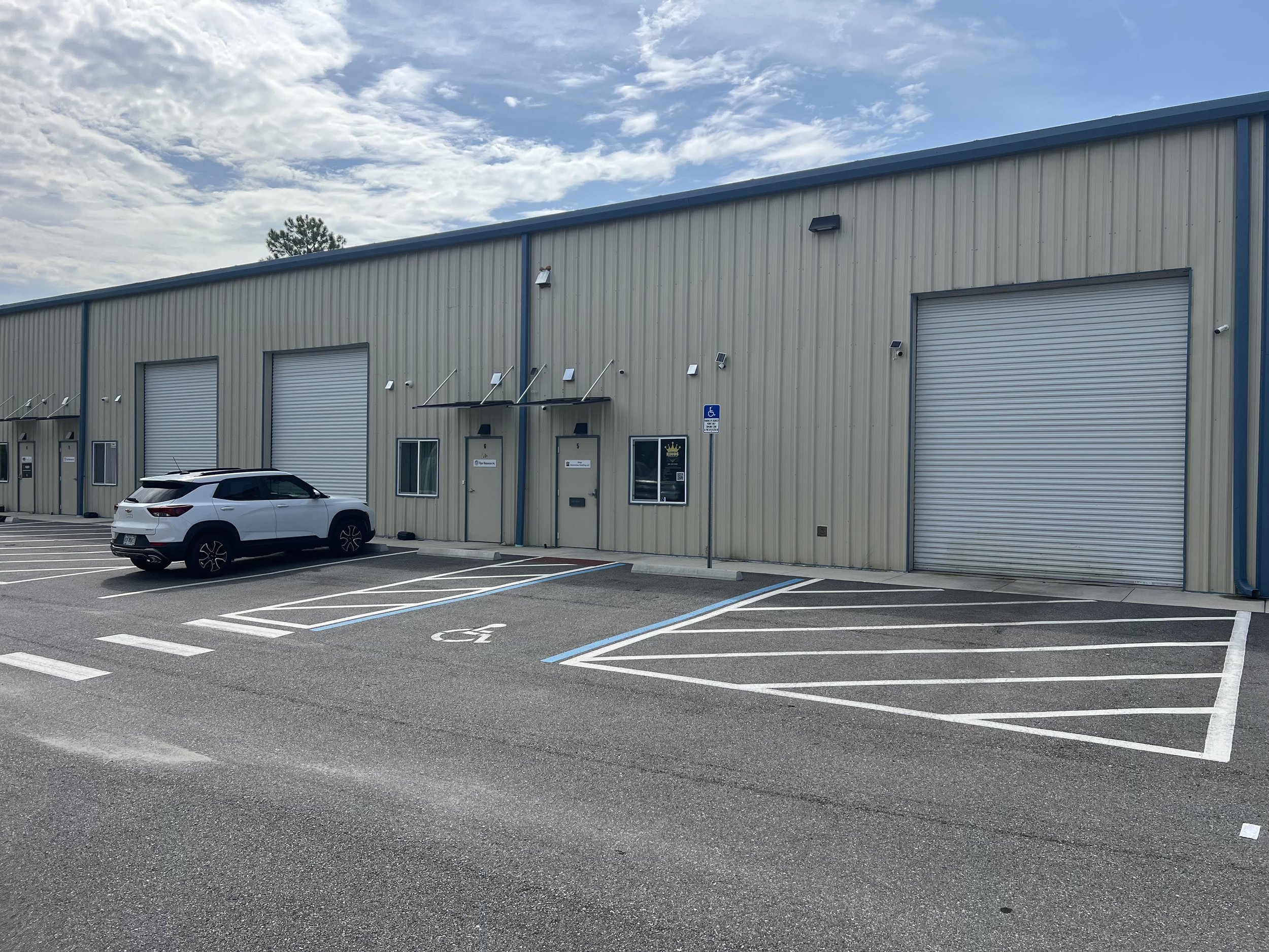 A parking lot outside a large industrial building with a beige metal exterior and closed roll-up doors. There is a white vehicle parked in a designated space, a handicap parking sign, and a few street lamps. The sky above is partly cloudy.