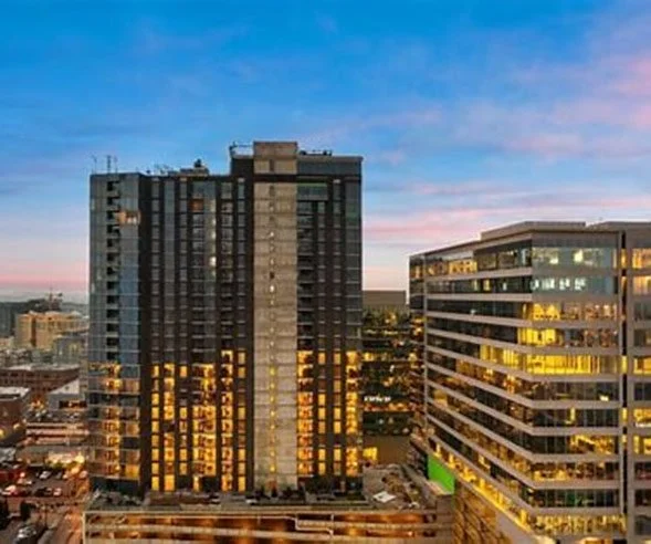 Cityscape with tall buildings at dusk, some windows illuminated, with a colorful sky in the background.