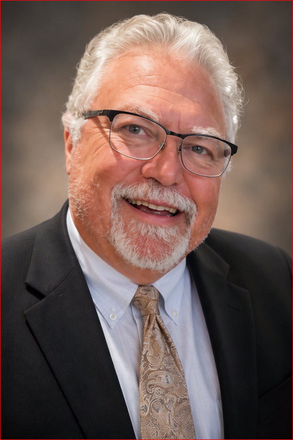 A smiling older man with white hair, a beard, and glasses, wearing a black suit, white shirt, and patterned tie.