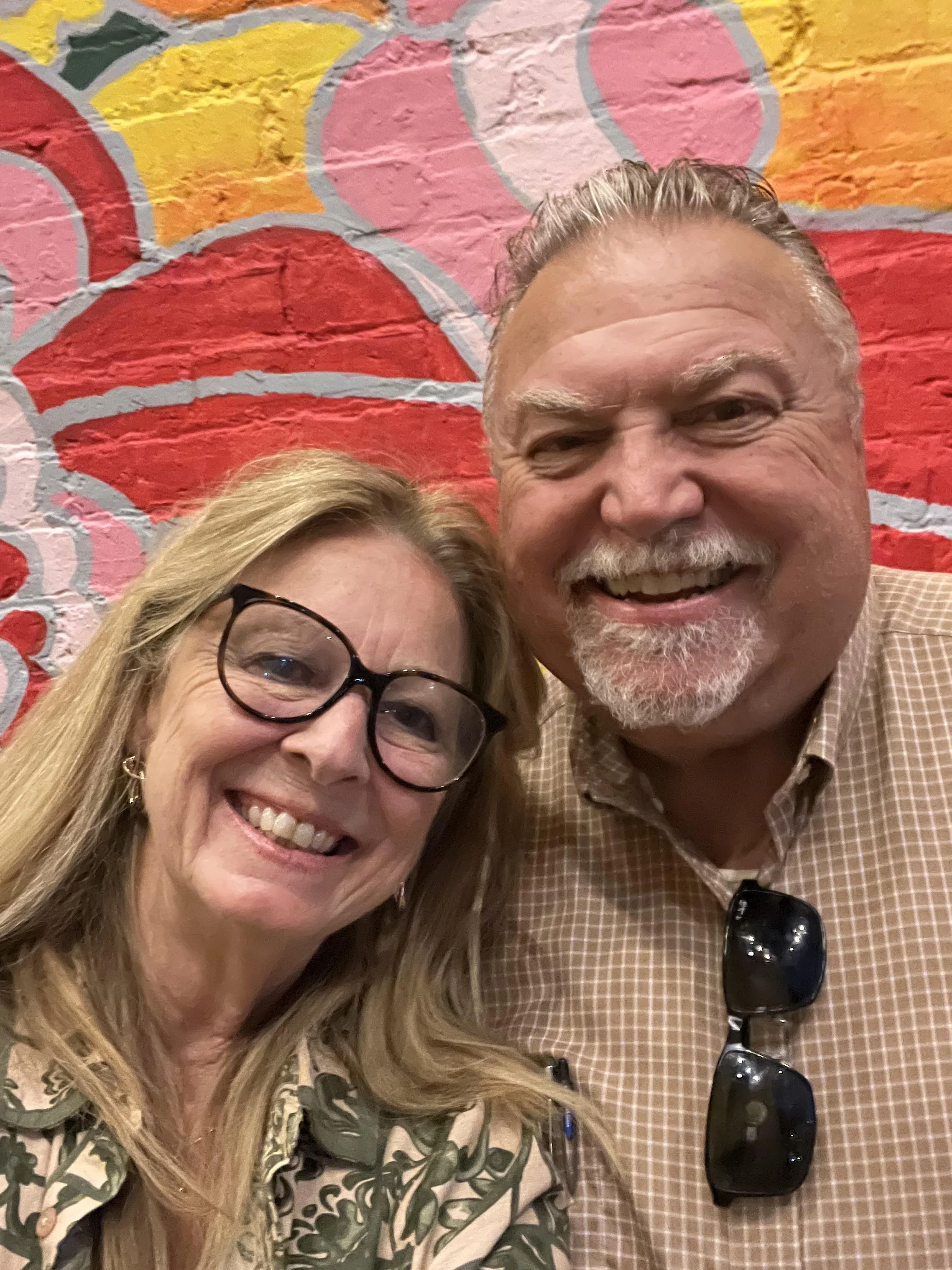 A smiling woman with glasses and blonde hair, and a smiling man with a beard and mustache, takes a selfie in front of a colorful painted brick wall.