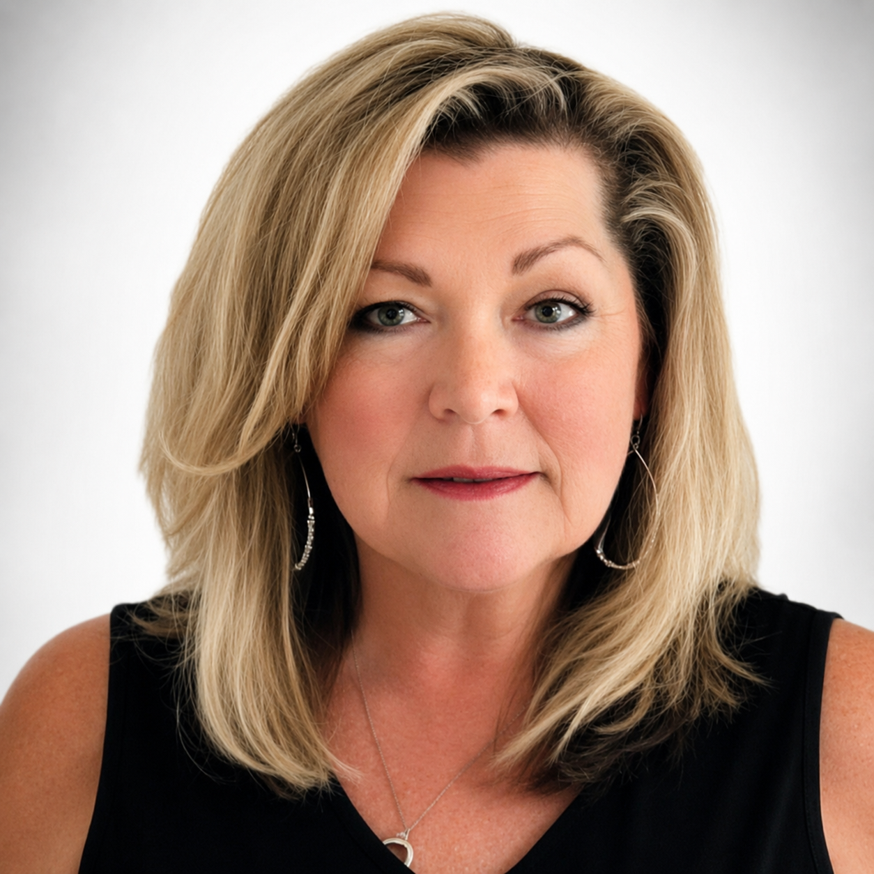 Close-up portrait of a middle-aged woman with blonde hair, wearing earrings, a necklace, and a sleeveless black top, looking directly at the camera with a neutral expression.