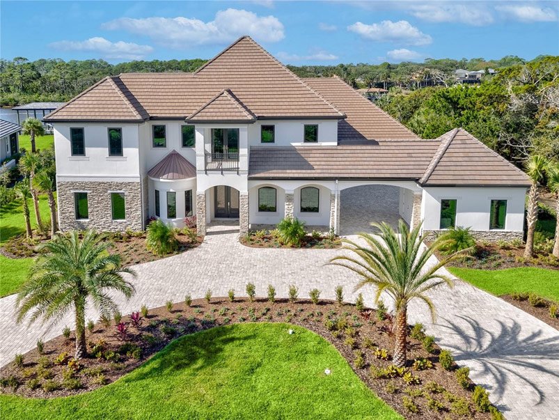 Large two-story house with stone and white exterior, surrounded by landscaped yard with palm trees and a paved driveway.