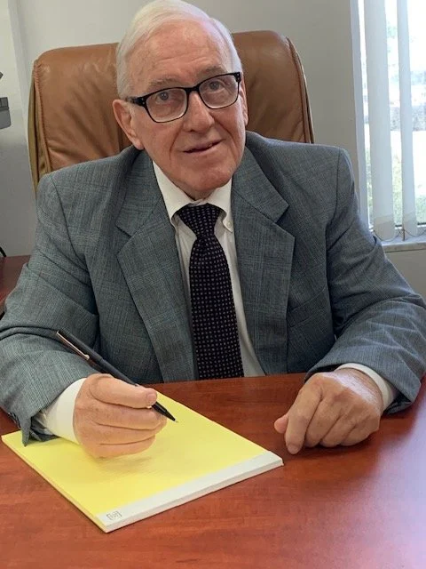 An elderly man wearing glasses, a gray suit, and a black tie, sitting at a wooden desk with a yellow notepad and a pen, in an office setting.