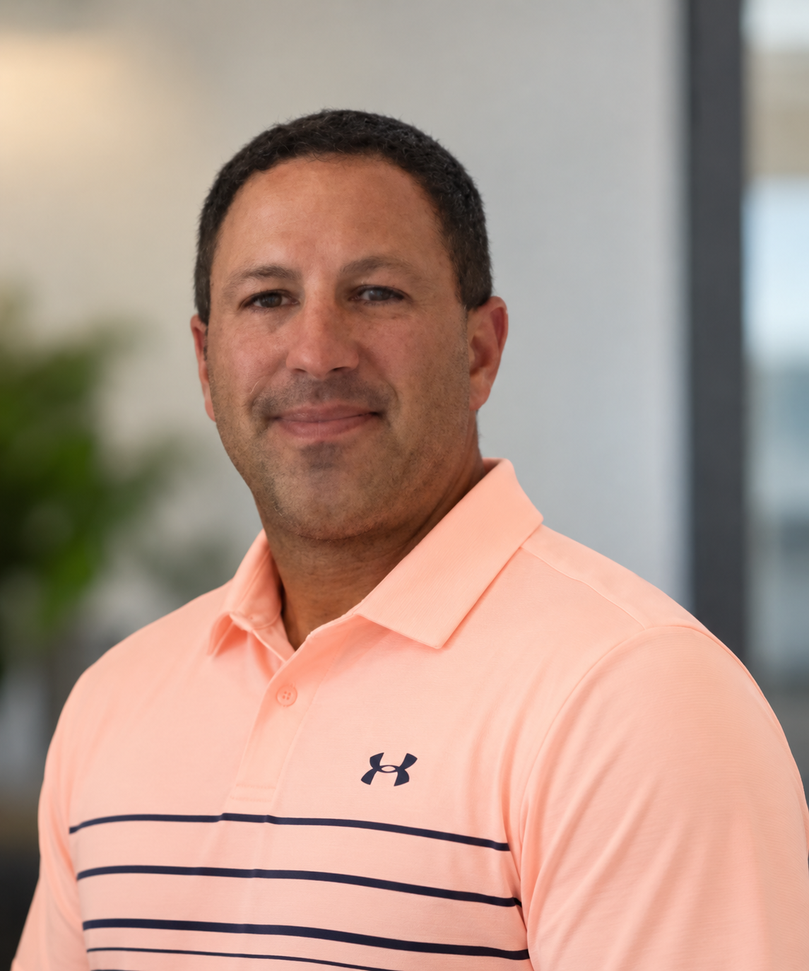 A man with short dark hair, wearing a peach Under Armour polo shirt with thin navy horizontal stripes, smiling slightly, in a modern indoor setting.