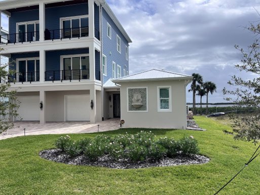 Front yard of a large blue house with white trim, showing a smaller white structure and a landscaped garden bed with grass and flowers, in a coastal area with palm trees and water in the background.