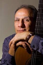 A man holding an acoustic guitar, resting his chin on his hand, looking at the camera.