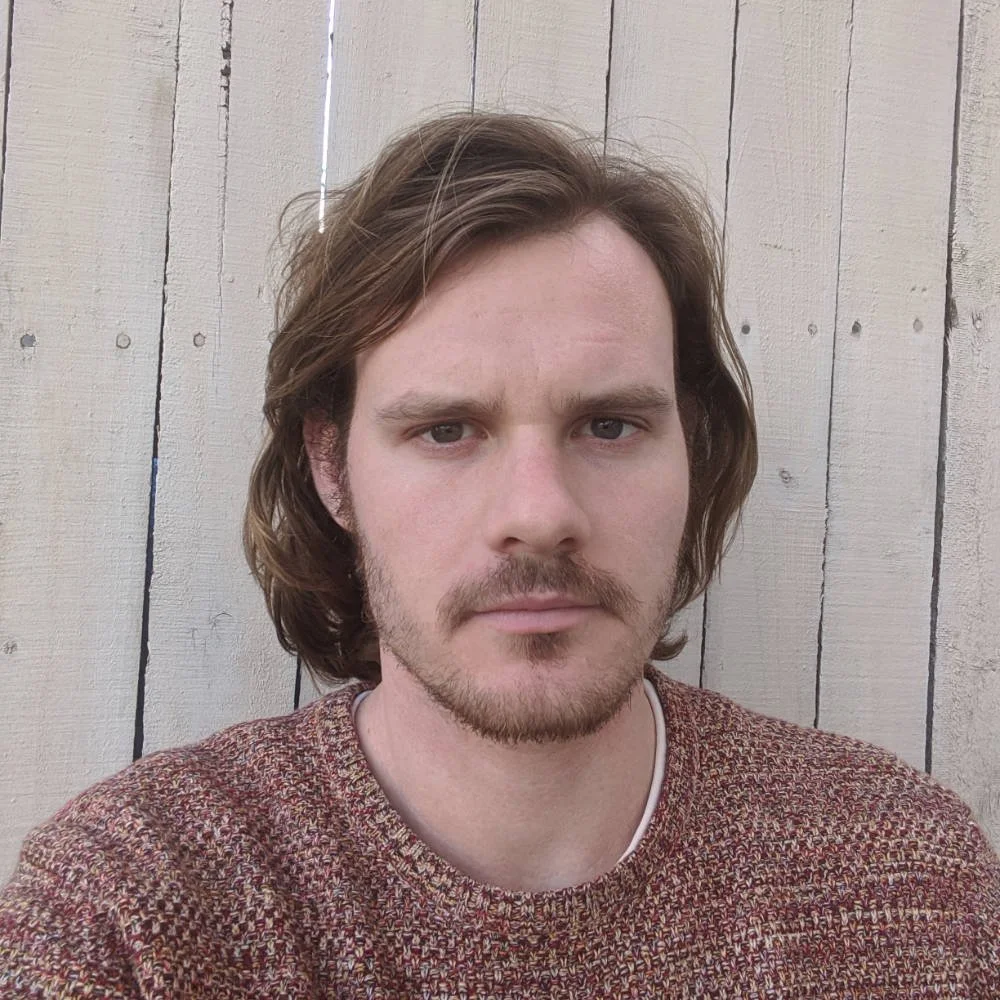 A young man with shoulder-length brown hair, facial hair, and wearing a patterned maroon sweater, taking a selfie in front of a light-colored wooden fence.