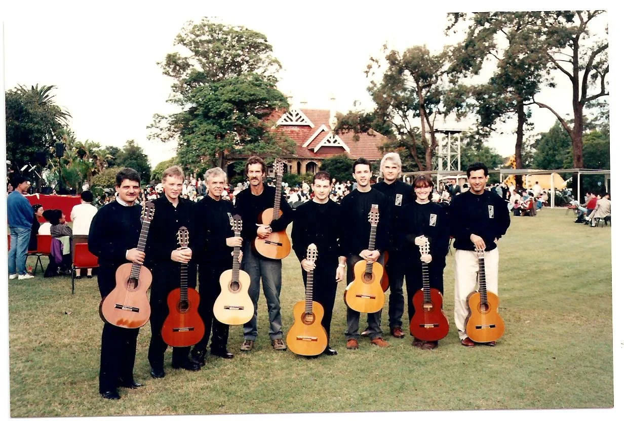 Group of nine people holding guitars, standing outdoors on a grassy area during an event, with trees, a house-like structure, and people in the background.