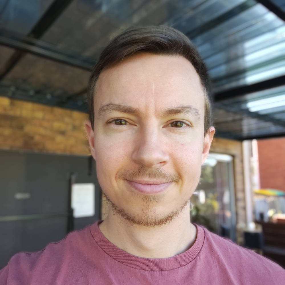 A young man with light skin, short dark brown hair, and a light beard, smiling outdoors on a sunny day, wearing a maroon T-shirt.