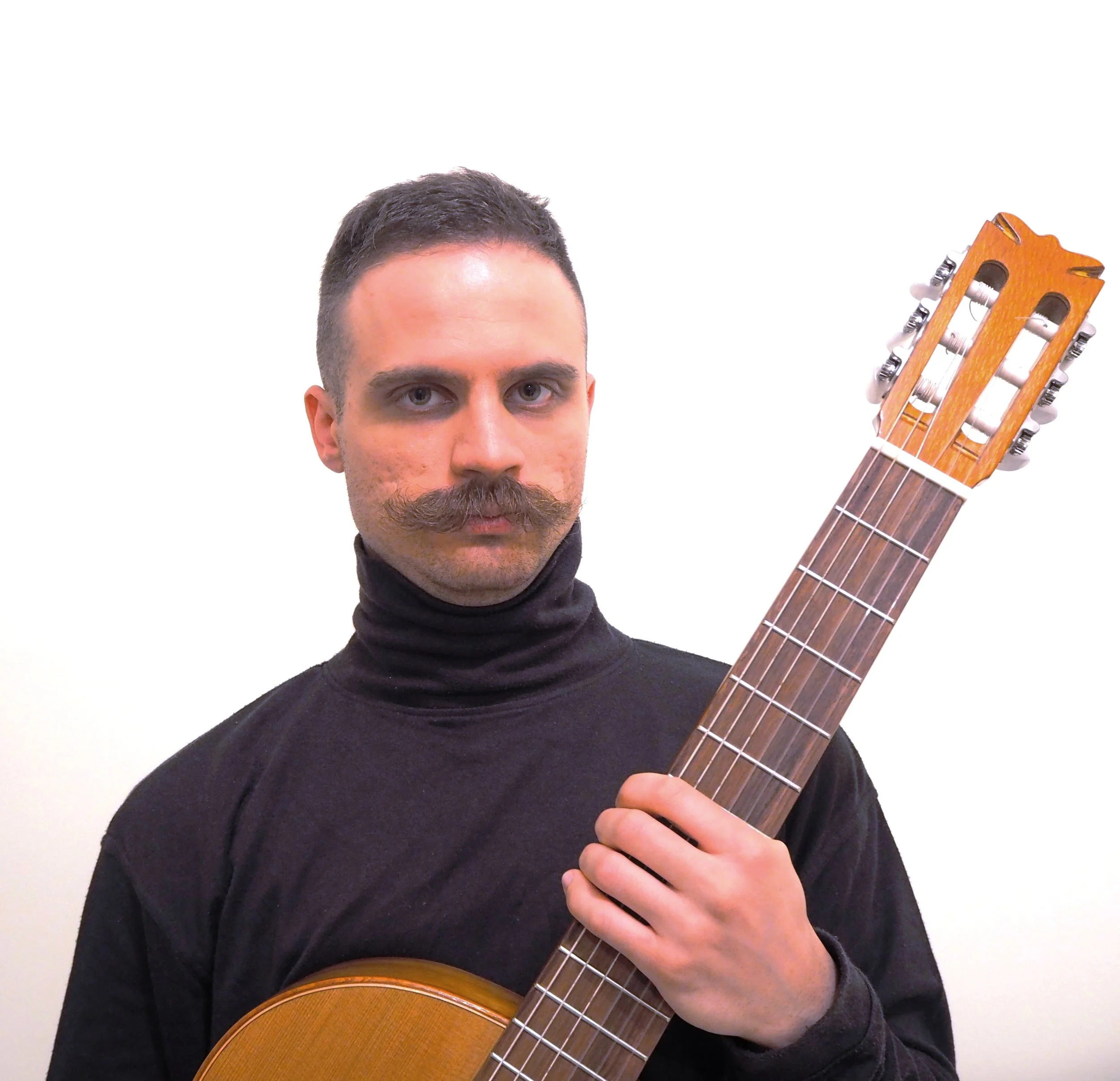 A man with short dark hair, a beard, and a mustache holding an acoustic guitar against a plain white background.