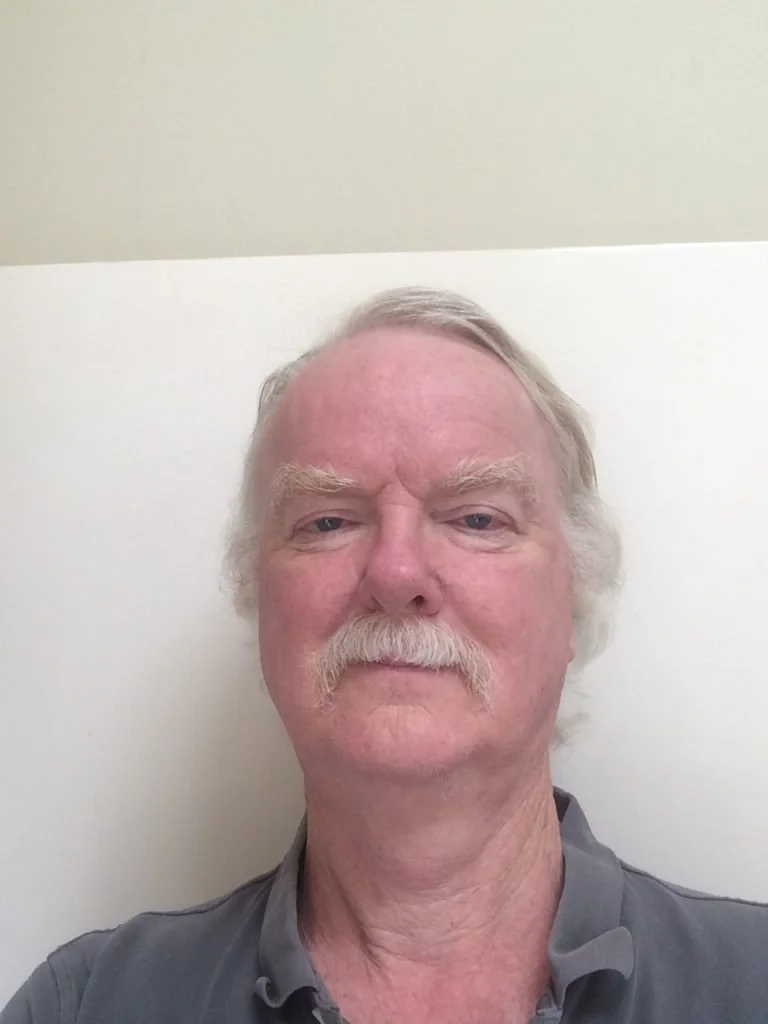 An older man with white hair, a white mustache, and a light complexion. He is wearing a gray collared shirt and standing against a background with a beige upper wall and a white lower wall.