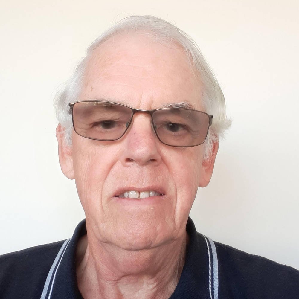 An elderly man with gray hair and wearing sunglasses, a dark collared shirt with white piping, standing against a plain white background.