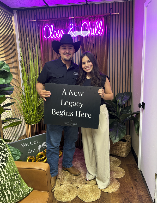 A man and woman standing together holding a sign that reads 'A New Legacy Begins Here.' The man is wearing a black cowboy hat and a black shirt, while the woman is dressed in light-colored pants and a dark top. Behind them is a neon sign that says 'Close & Chill,' along with green plants and a wooden wall.
