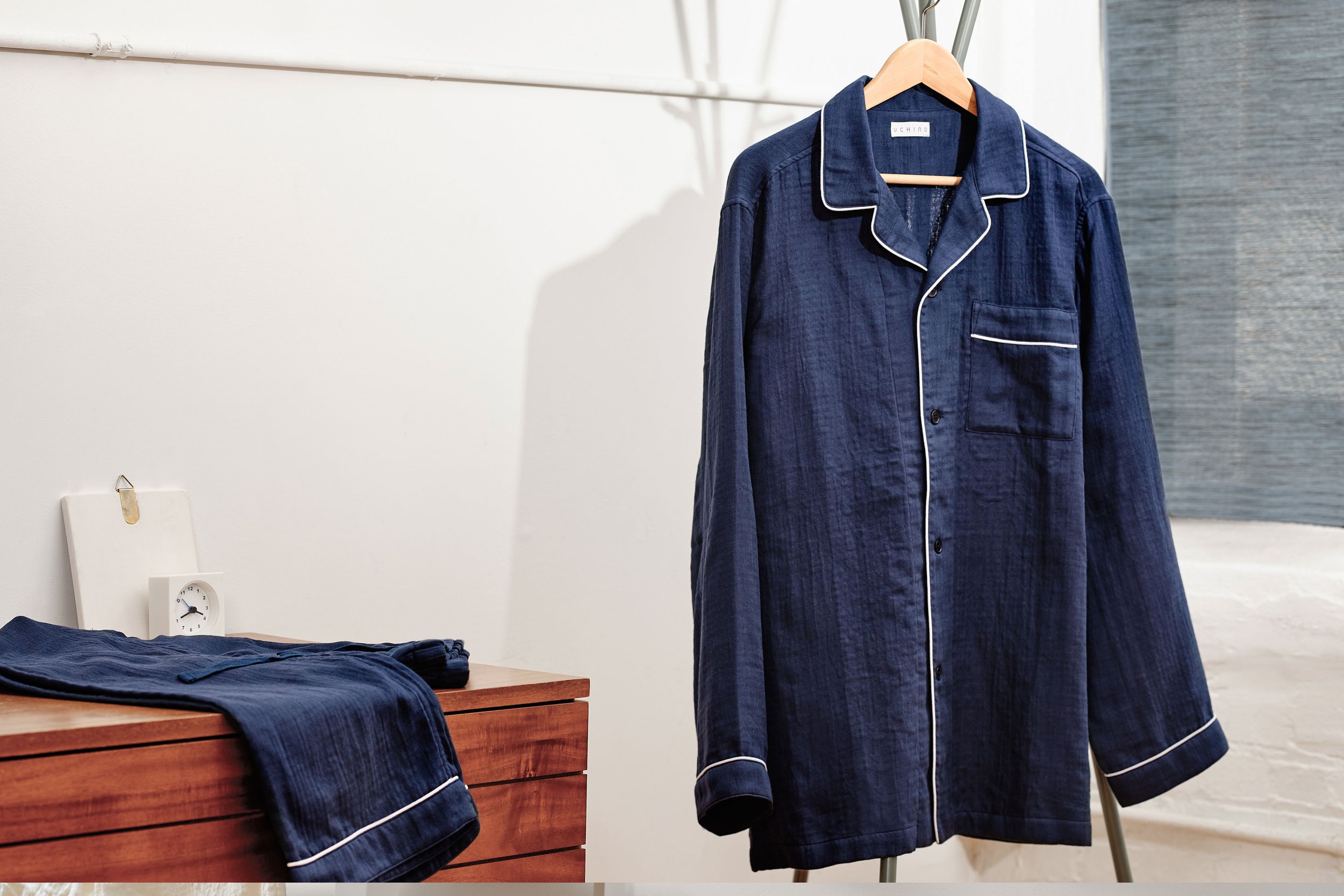 Marshmallow Gauze Pajama Navy Color. Pajama set with white piping hangs on a wooden hanger. The set includes a long-sleeved shirt and matching pants, displayed in a minimalistic room with a wooden dresser, a small clock, and a white wall.