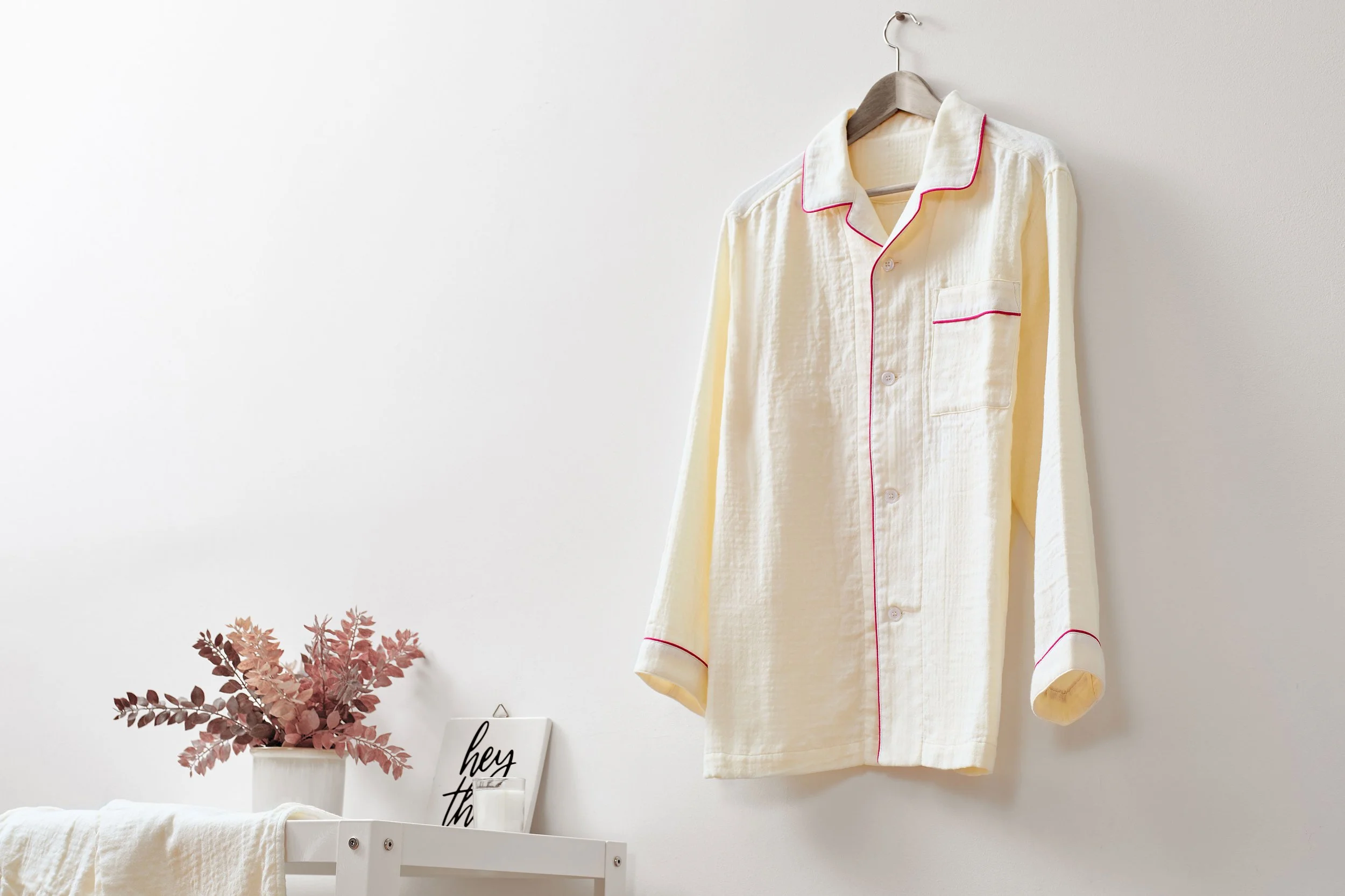Cream-colored UCHINO Marshmallow Gauze Pajama. A cream-colored pajama shirt with red piping hangs on a wooden hanger against a white wall.