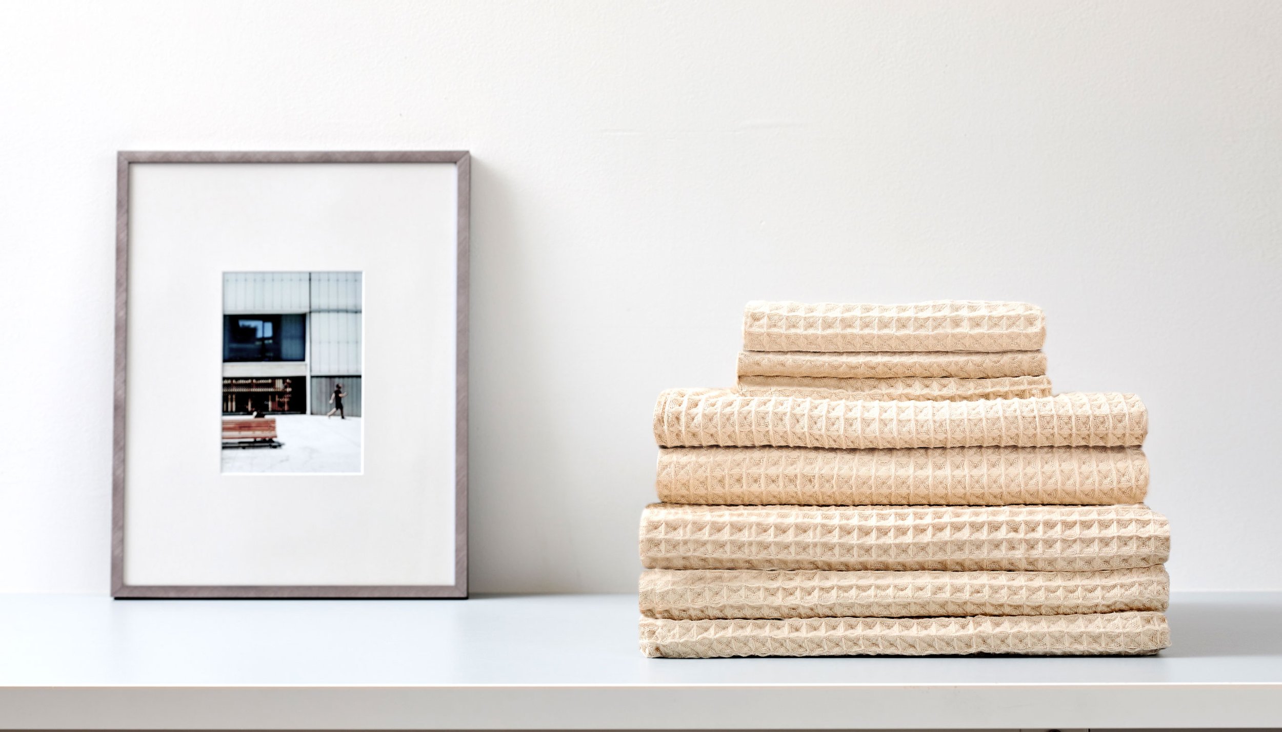 Stacked beige textured towels on a white shelf next to a large framed photograph of a modern building with a person walking in front.