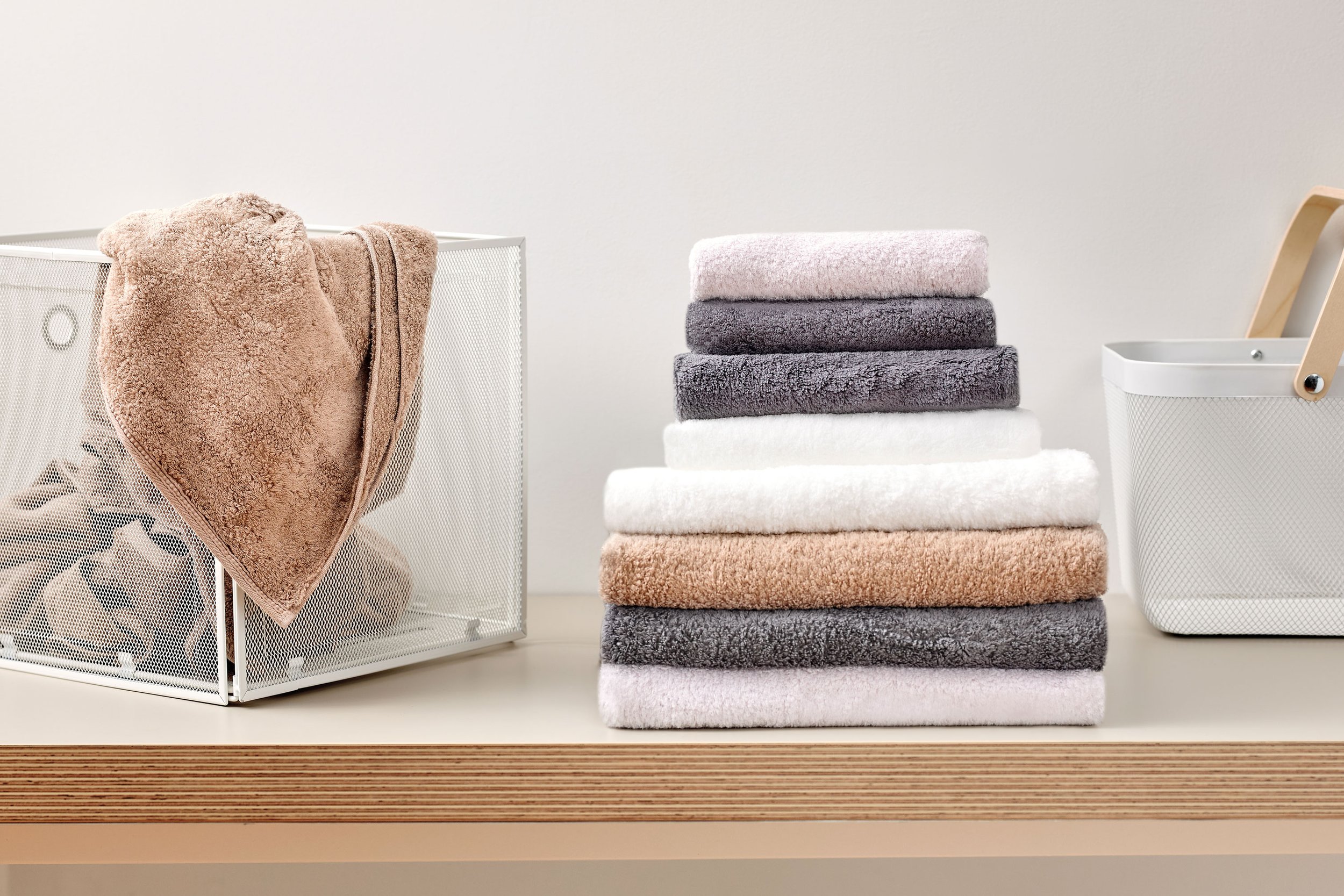 UCHINO Blissful Towels beige gray and white. To the left, there is a white wire basket with a beige towel draped over the edge. To the right, there is a white mesh container with a light wood handle, set against a plain off-white background.