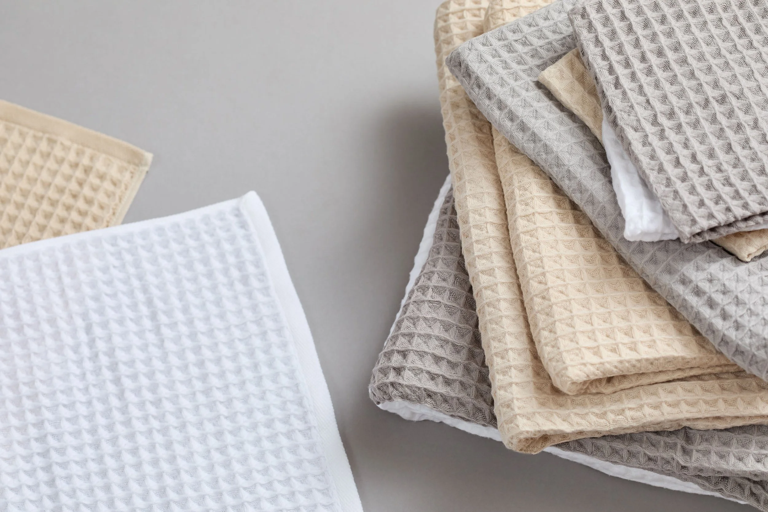 UCHINO Air Waffle towels. Stacked textured waffle-patterned towels in neutral colors, beige to light gray, on a gray surface with a plain background.