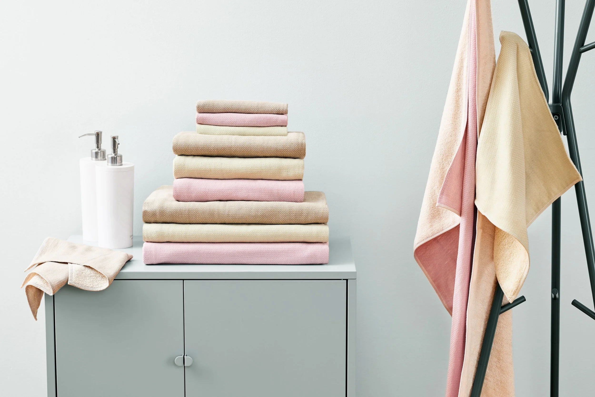 Stacked towels in pastel pink, beige, and cream colors on a light grey cabinet with a towel rack holding similarly colored towels to the right. Two white soap dispensers with silver pumps are to the left of the towels.