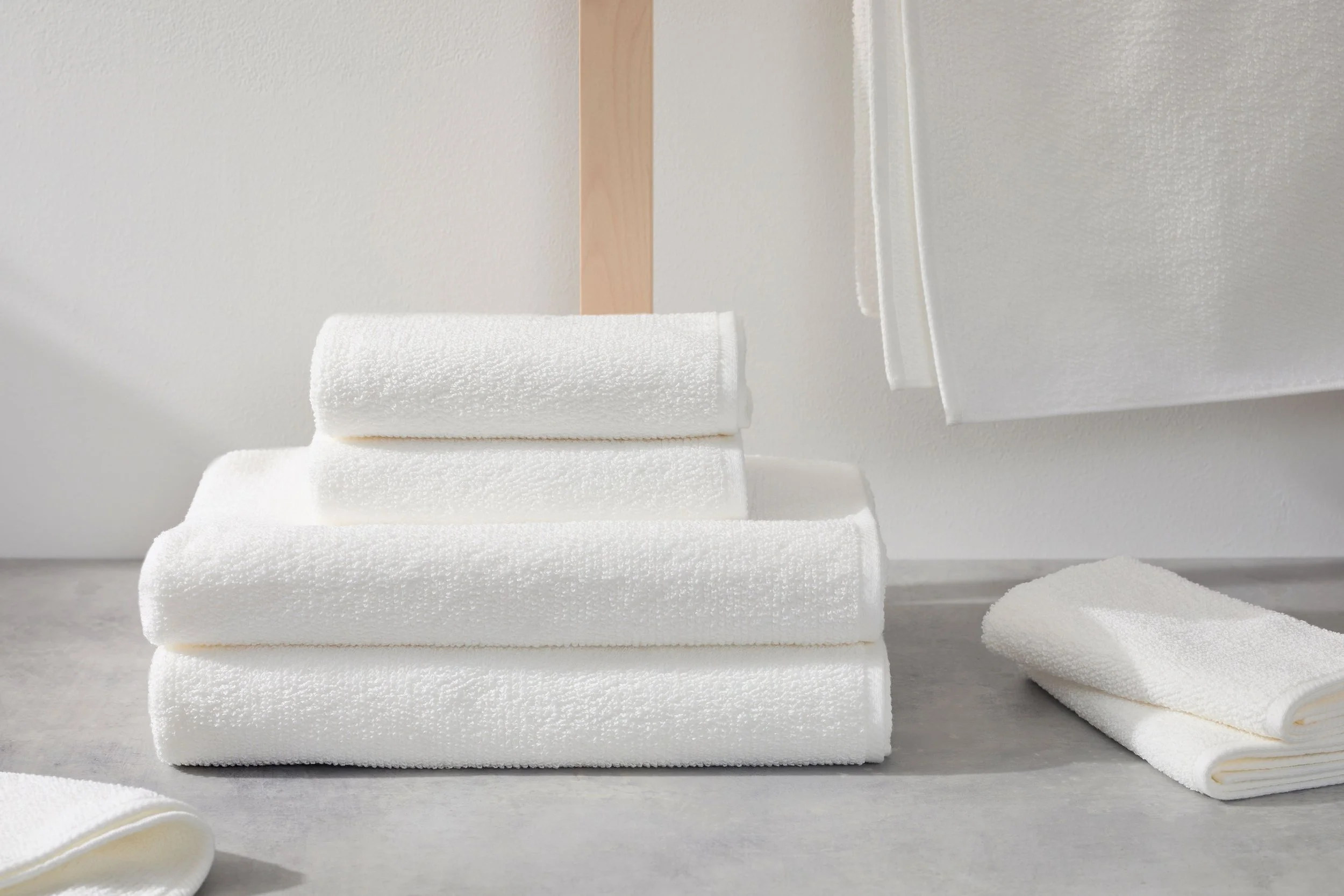 UCHINO blissful towels. Stacked white towels on a light gray surface with roll towels nearby and a white textured wall in the background.