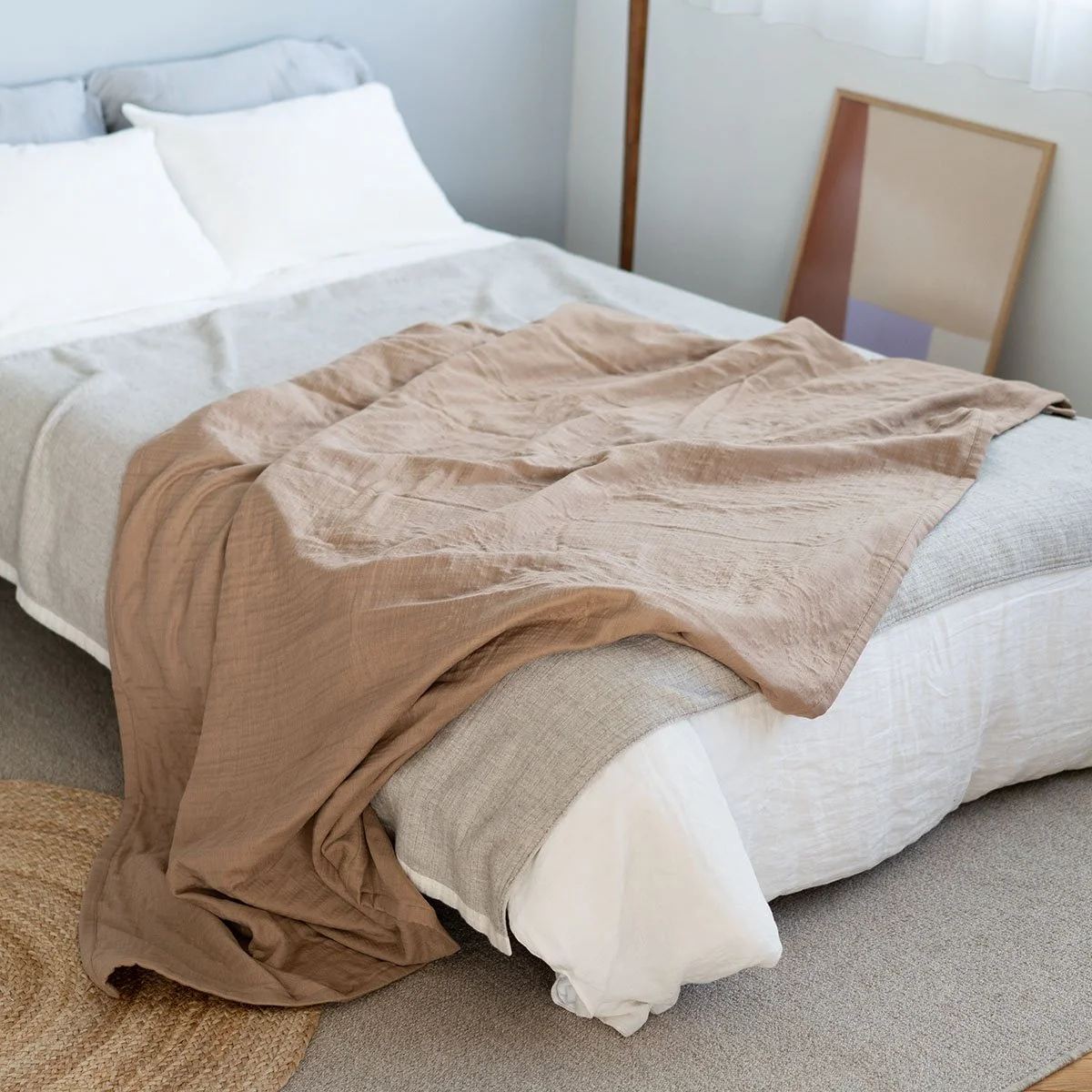 A neatly made bed with white pillows, a light gray blanket, and a beige bedspread, in a bedroom with light-colored walls, a window with sheer curtains, and a framed artwork leaning against the wall.