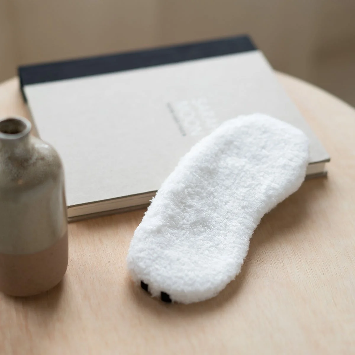White fluffy slipper on a wooden surface, with a beige ceramic bottle and a closed white notebook in the background.