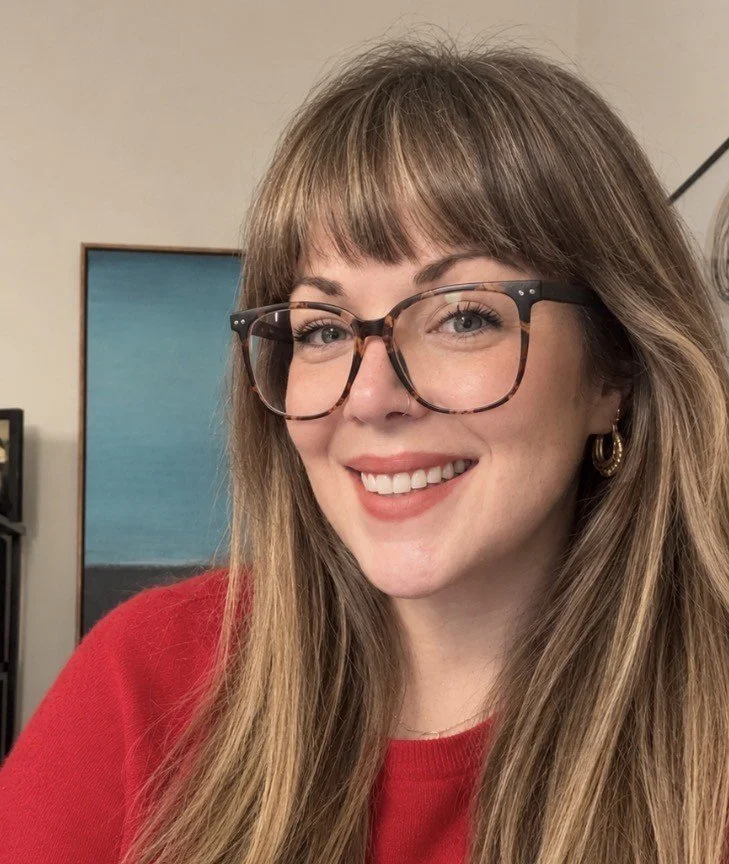 Woman in glasses and a red sweater smiling, sitting at a desk.