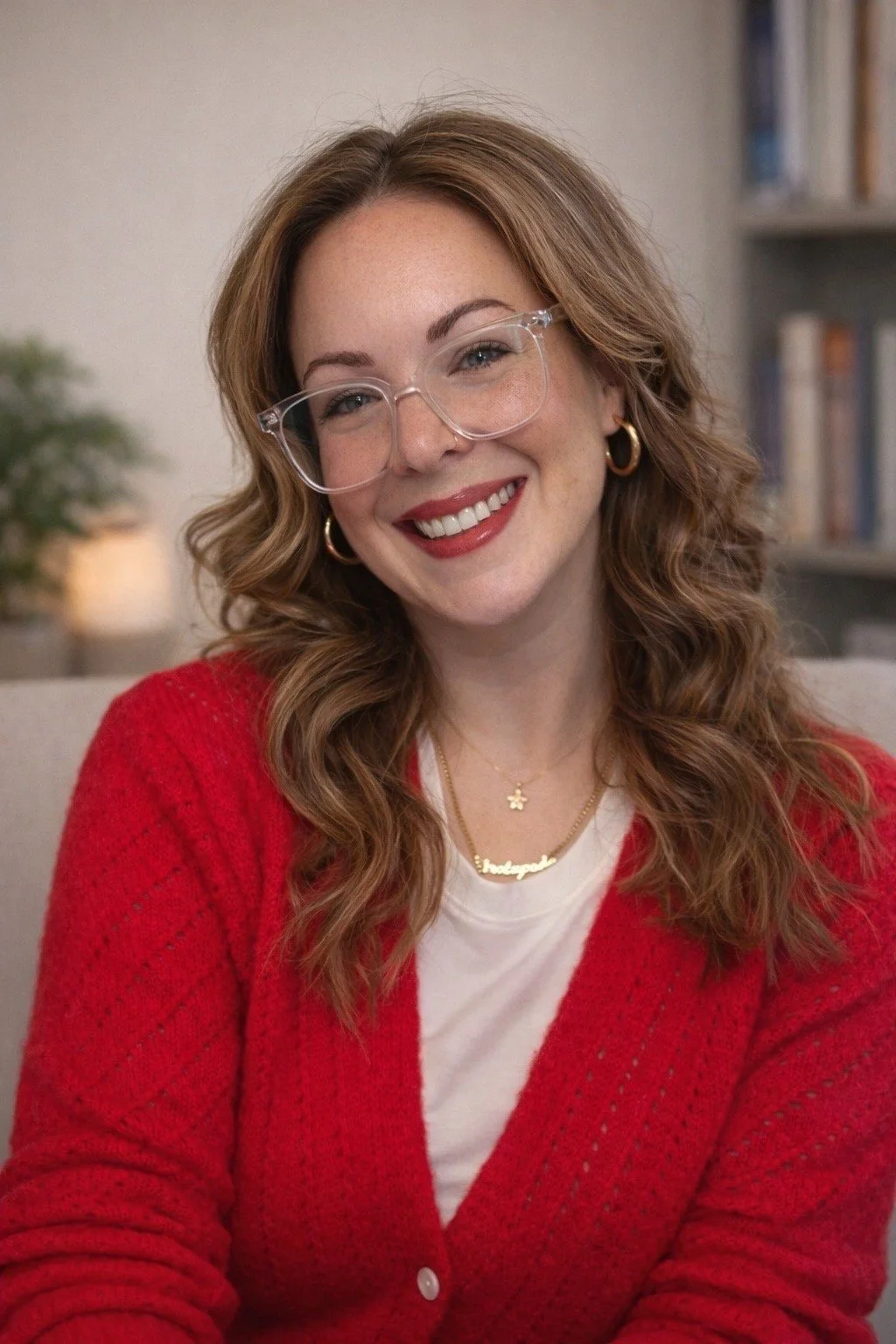 A smiling woman with curly red hair and glasses, wearing a red cardigan and a necklace, sitting indoors with a bookshelf in the background.