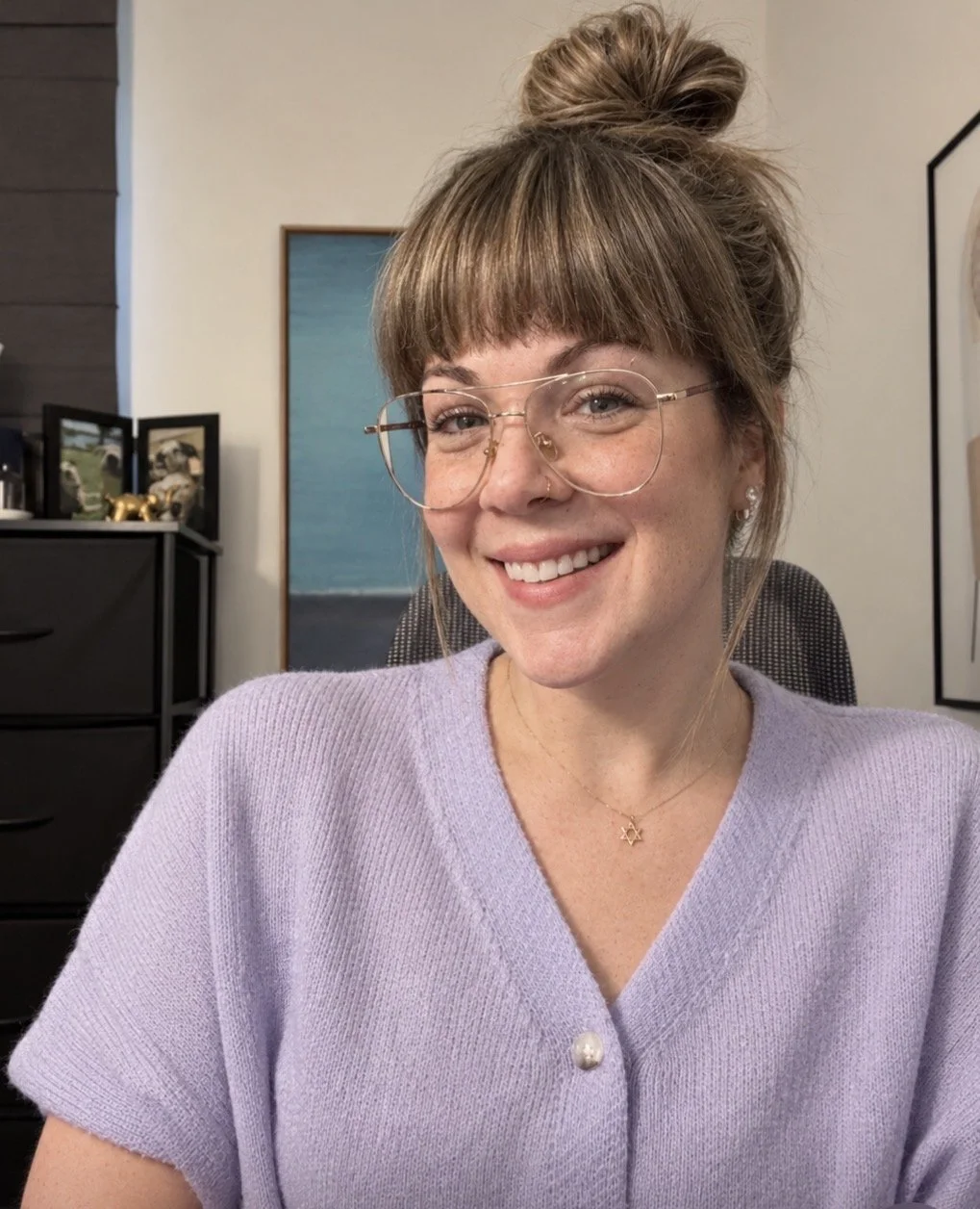Woman in her hair up, wearing glasses and smiling as she sits at a desk.