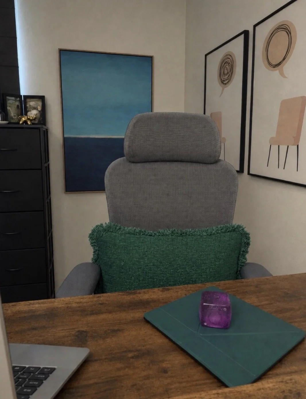A desk with a laptop, a green mouse pad, and a purple mouse. Behind the desk, there is a gray office chair with a green pillow. The background features framed artwork on the wall, including abstract depictions of chairs and circular designs.