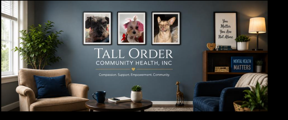 Interior of an office or community space with a blue wall featuring framed pictures of dogs, a tall green plant, and a sign that says 'Tall Order Community Health, Inc.' with the slogan 'Compassion. Support. Empowerment. Community.' A beige armchair with a navy cushion, a side table with a mug and book, a dark blue armchair, a wooden shelf with books and a potted plant, a lamp, and decorations including a wooden heart and a giraffe figurine.