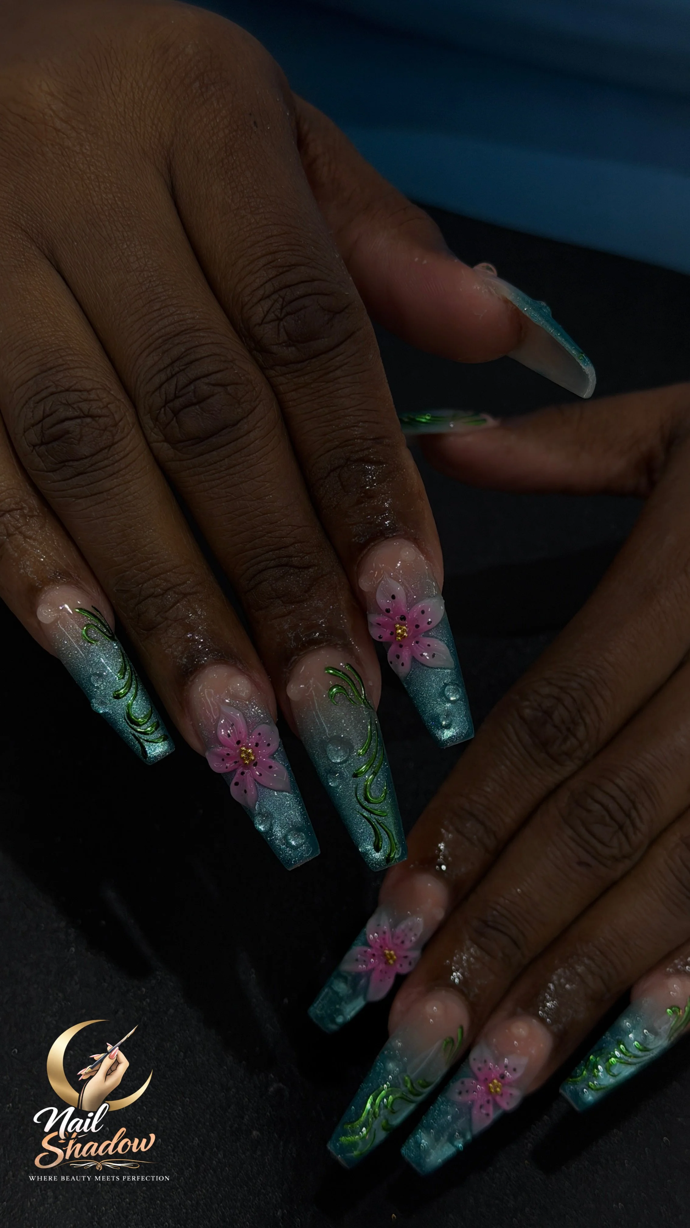 Close-up of a person’s hand with long, decorated acrylic nails featuring pink flowers, green leaves, and shimmering blue accents.
