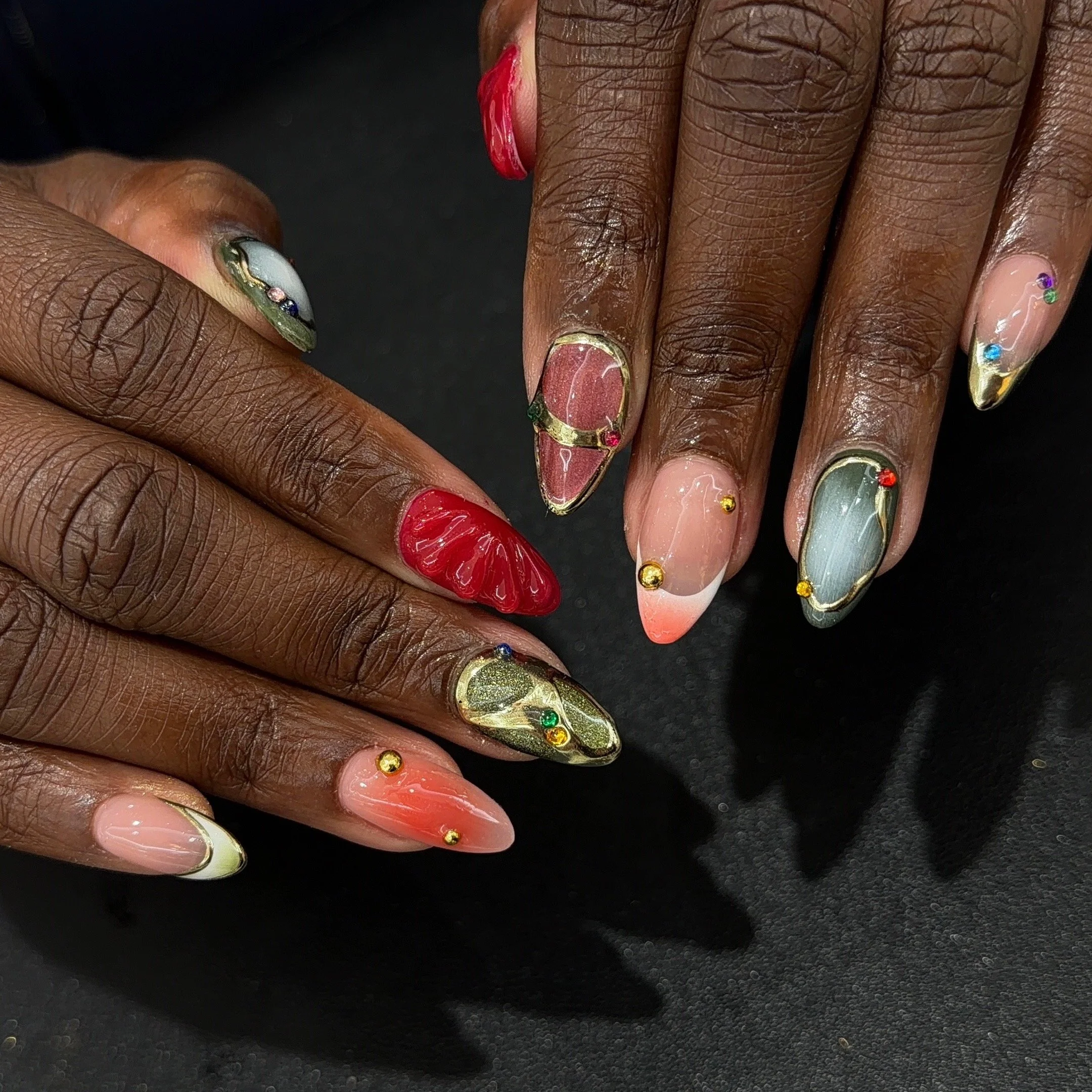 Close-up of two hands with decorated, colorful, floral and geometric nail art and small gold and multicolored rhinestones on long, pointed nails against a black background.