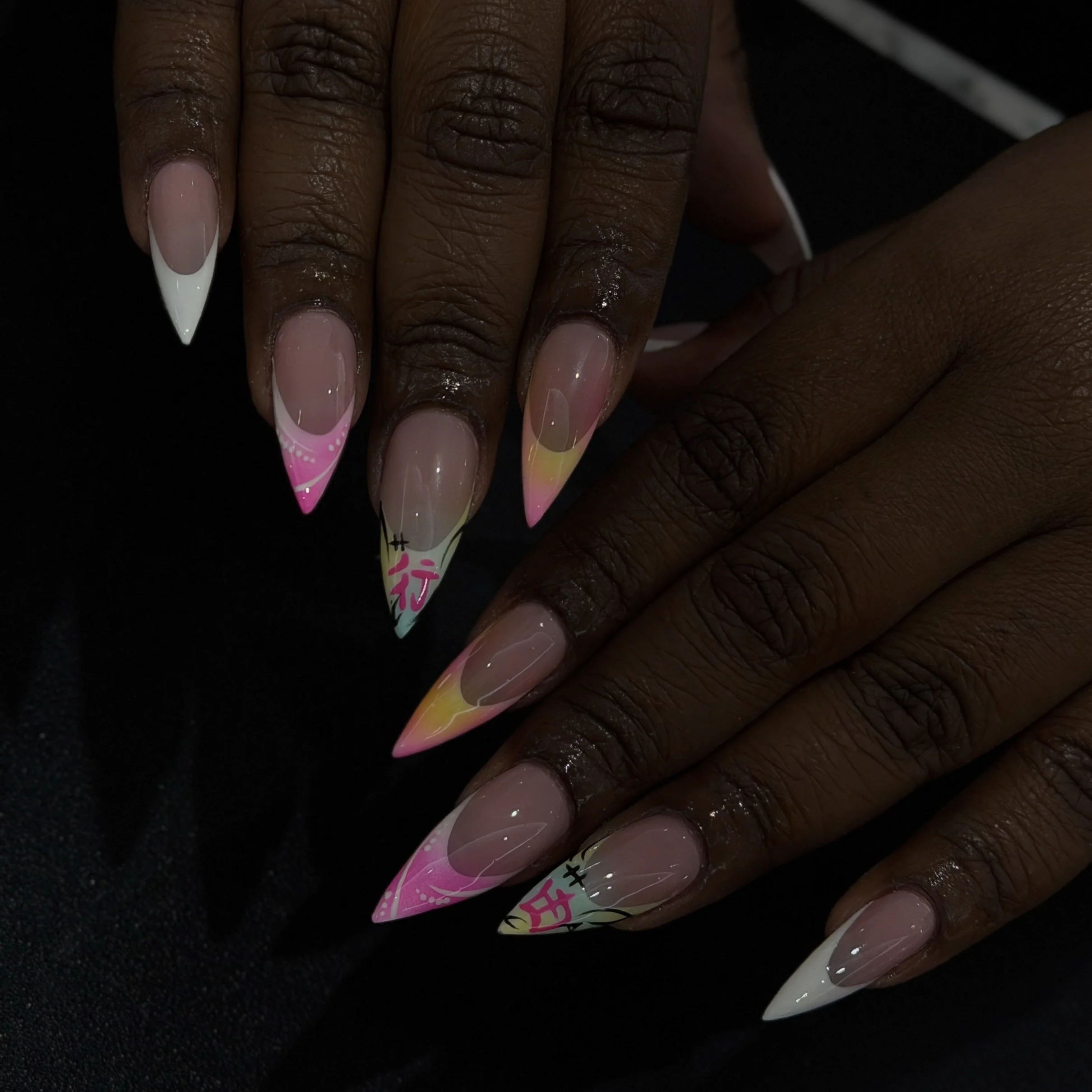 Close-up of a hand with long, decorated acrylic nails painted in pastel pink, yellow, and white with designs and text, against a black background.
