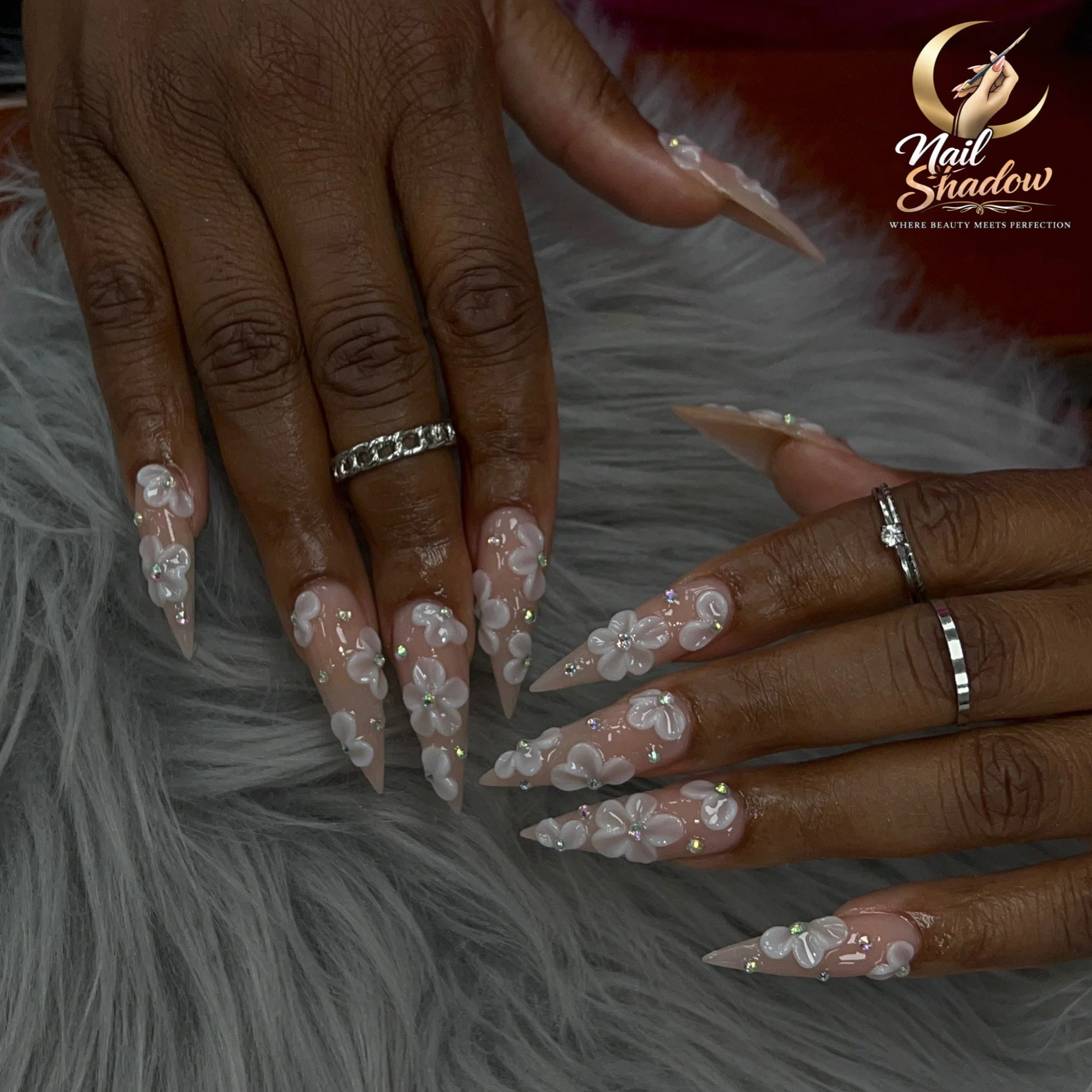 Close-up of a woman's hand with long, decorated nails, rings, and dark skin, resting on a gray furry surface. The woman is getting a manicure with her nails painted and embellished with rhinestones. There is a logo on the upper right corner that reads 'Nail Shadow' with a hand holding a brush and a tagline 'Where beauty meets perfection'.