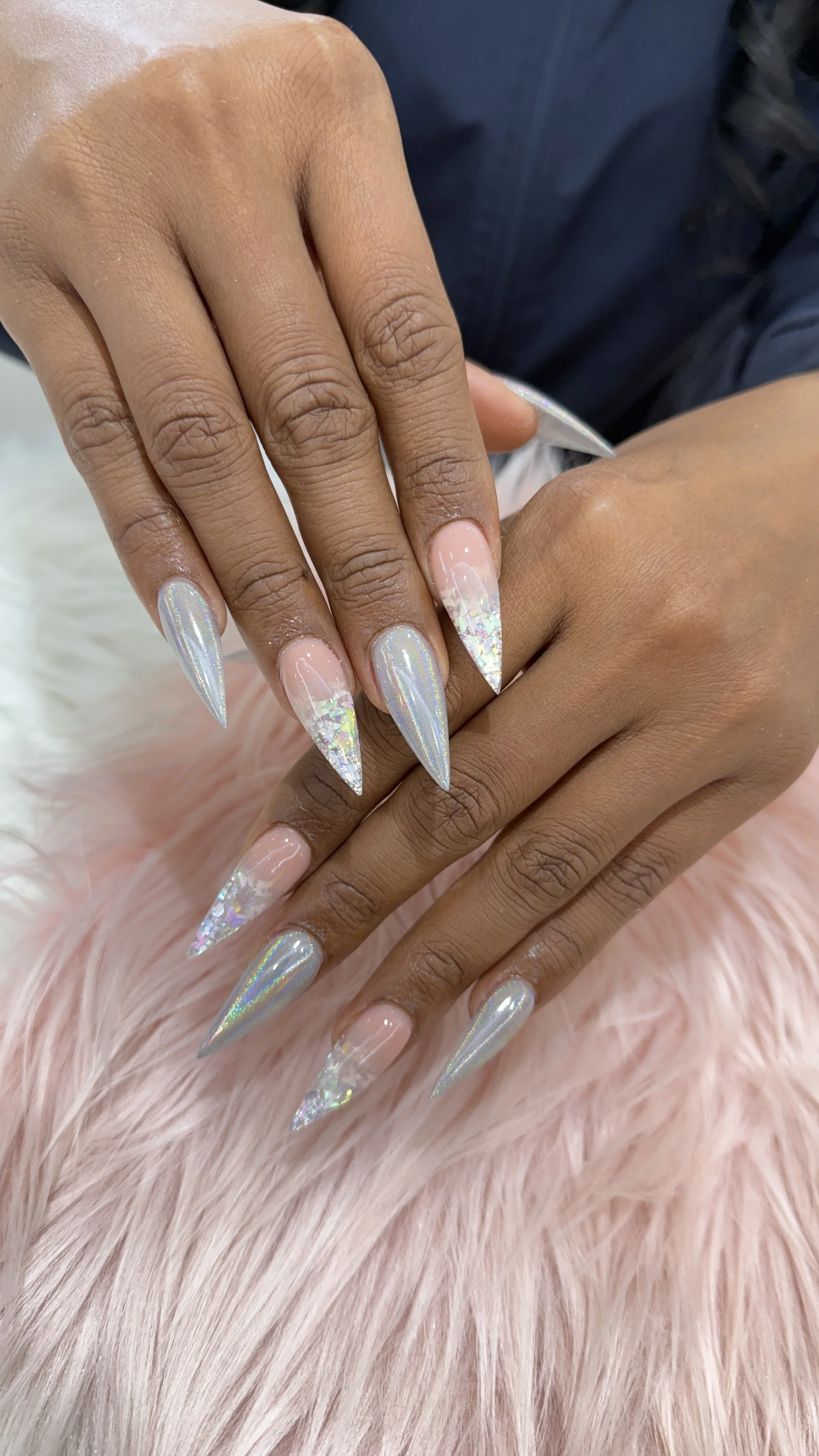 Close-up of a person's hand with long, pointed, holographic nails with iridescent glitter, resting on soft pink faux fur.