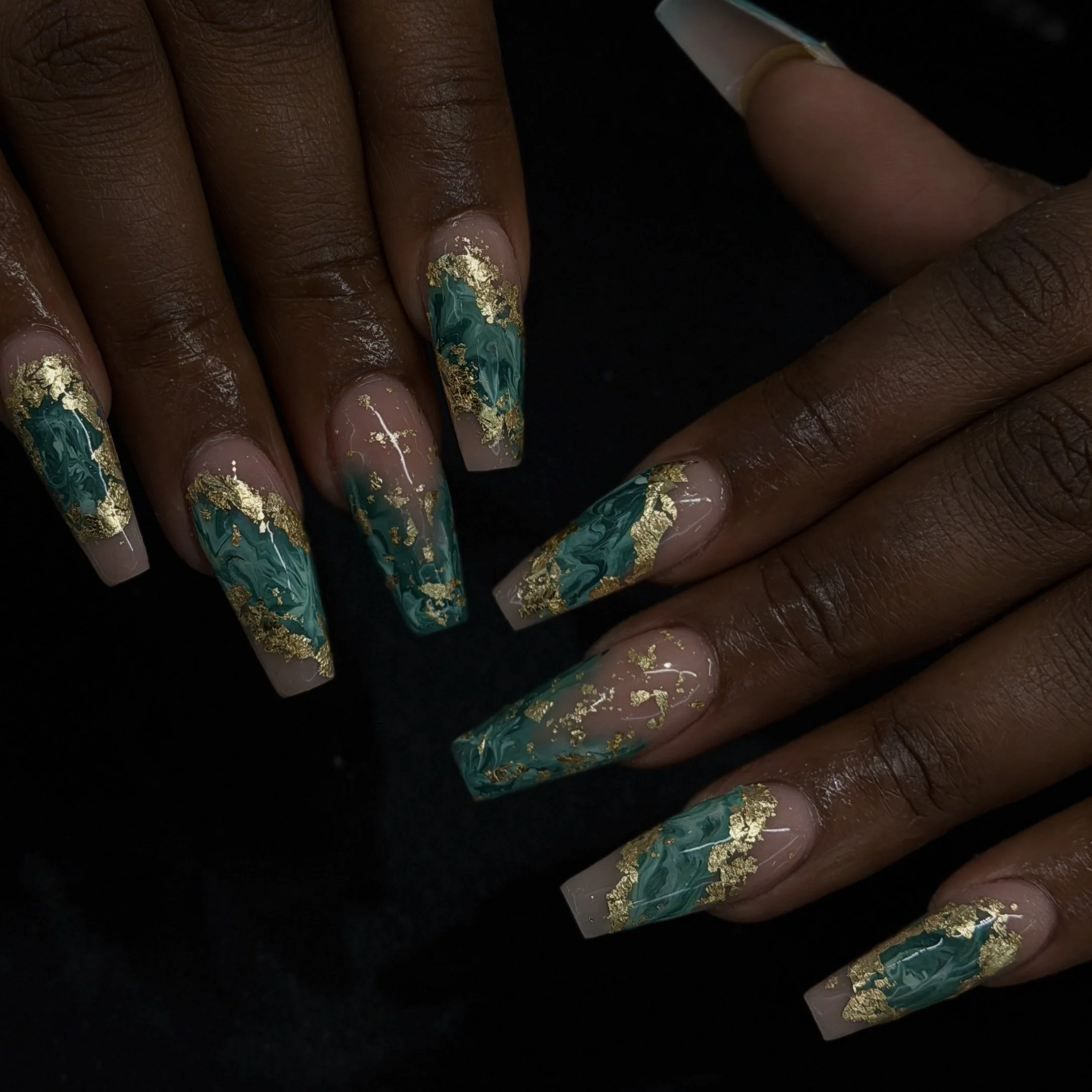Close-up of a hand with dark skin and decorated nails featuring turquoise, gold, and marble-like patterns, with a black background.