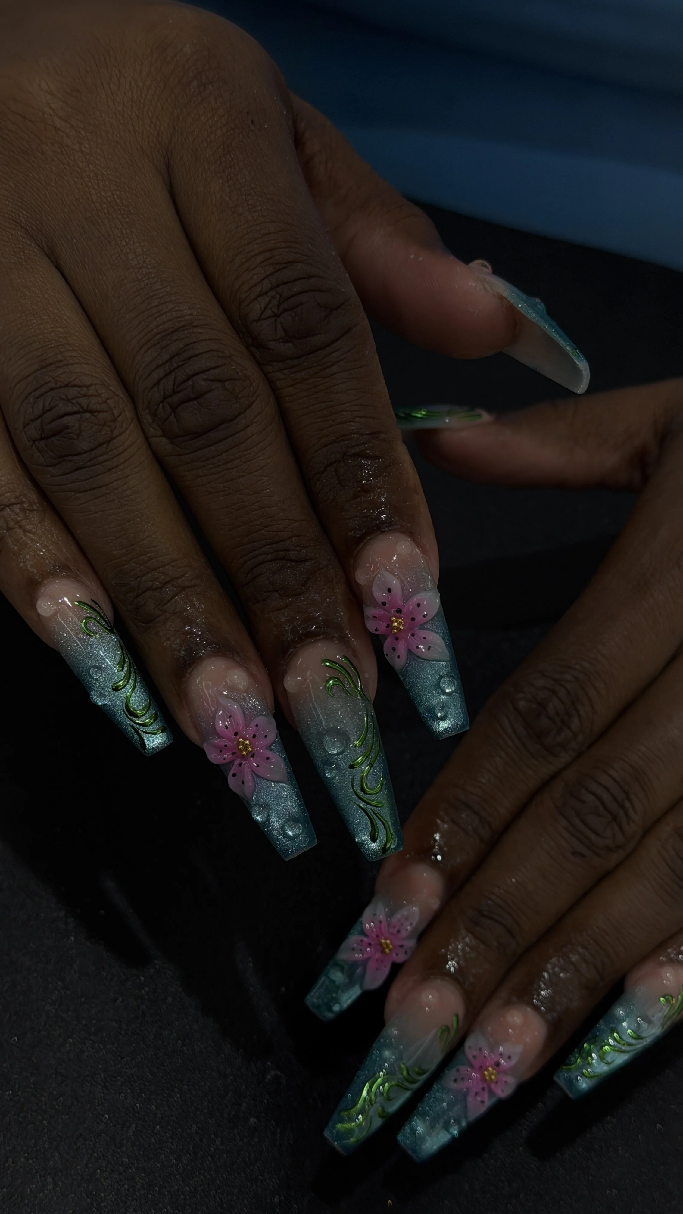 Nail art with pink flowers, green leaves, and blue glitter on long, square-shaped acrylic nails.