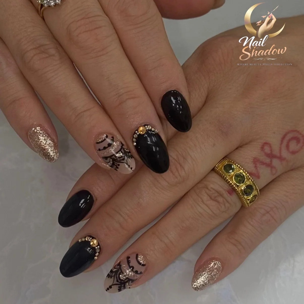 Close-up of manicured hands with decorated black, gold glitter, and floral nail designs, wearing a gold ring with green stones and a pink tattoo on the finger. The image features a "Nail Shadow" logo in the top right corner.