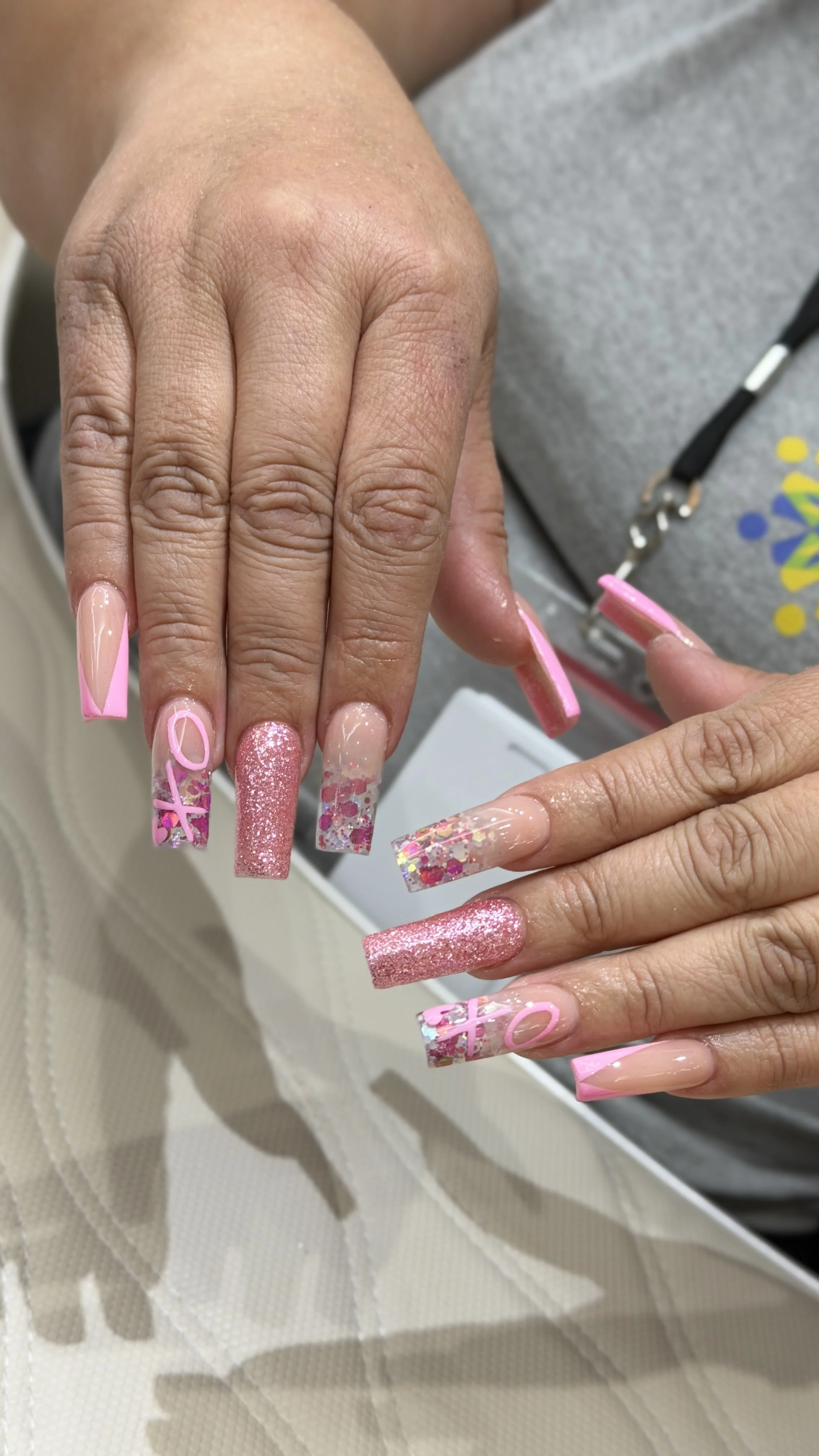 Close-up of a person with long, decorated pink and glittery acrylic nails, with some nails featuring the word 'LOVE' in pink and glitter, while the other nails have pink and glitter patterns. The person is holding their hand up, showing the nails cle