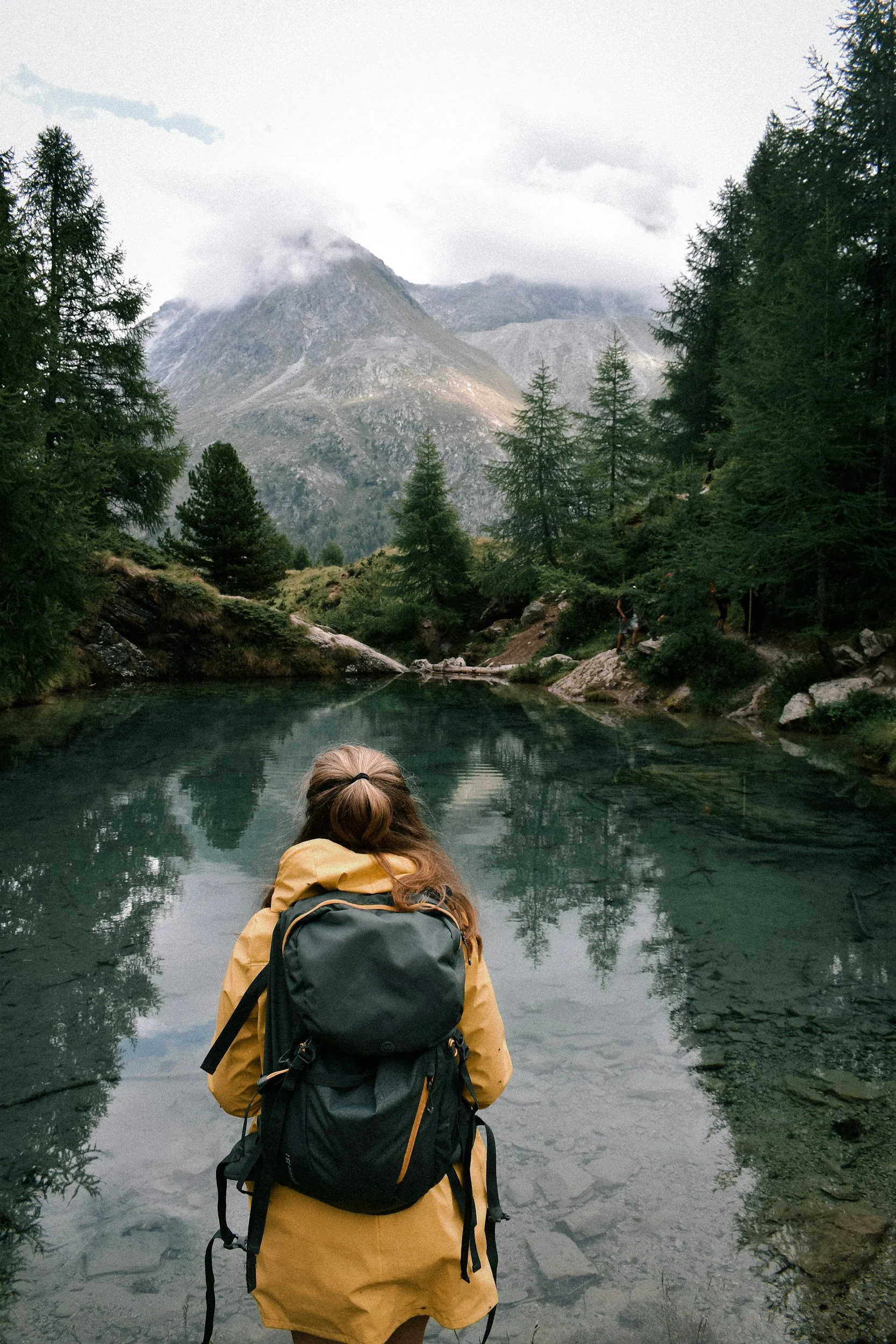 A person with a yellow rain jacket and a black backpack sitting by a river, surrounded by pine trees, with mountains and cloudy sky in the background.