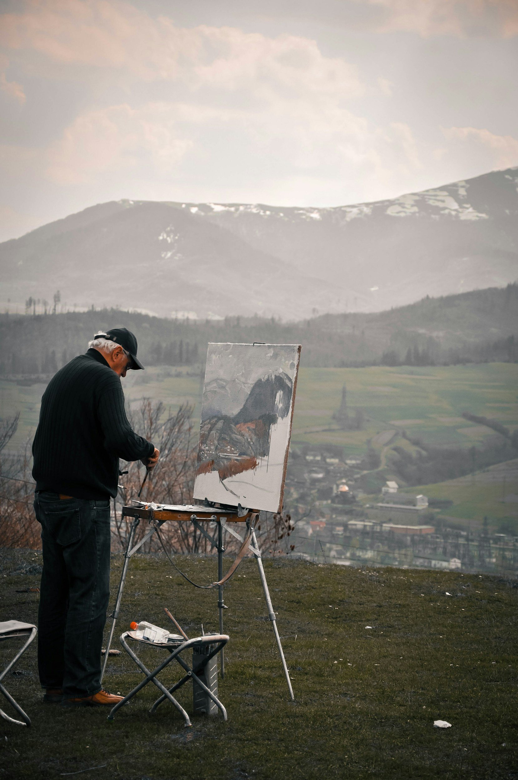 An elderly man with white hair and a black cap is painting outdoors on a canvas set on an easel. The scene includes a rural landscape with rolling hills, some snow patches, and a cloudy sky in the background. The man is wearing a black sweater and jeans, standing at an outdoor painting setup with a folding chair nearby.