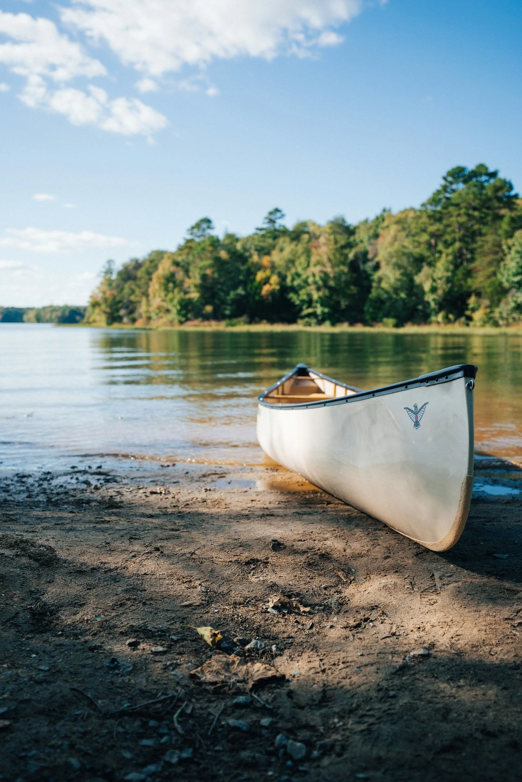 White canoe resting on a sandy shore beside a calm river, with a forested hill in the background under a partly cloudy sky.