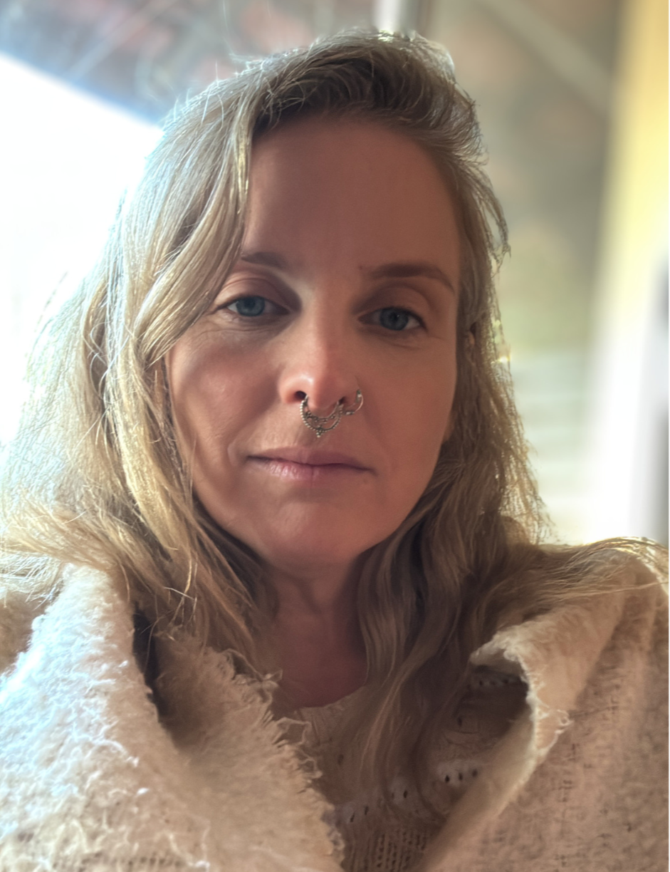 A woman with blonde wavy hair, blue eyes, and multiple nose piercings takes a close-up selfie indoors with a blurred background and soft lighting.