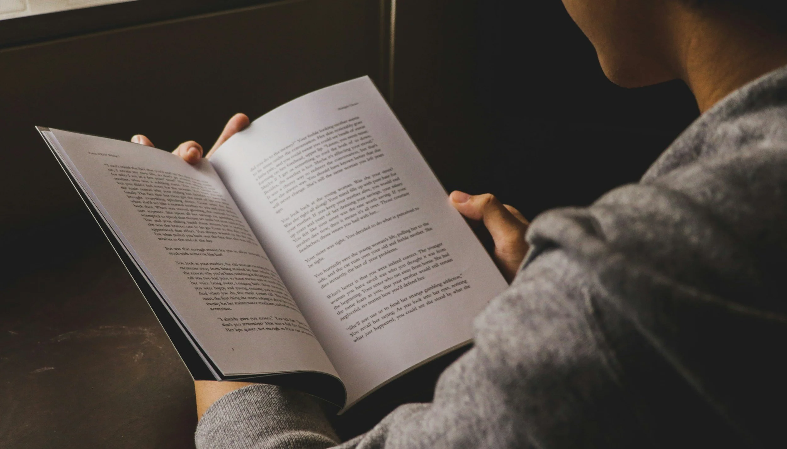 Person reading a book or document, wearing a gray long sleeve shirt, sitting at a dark surface.