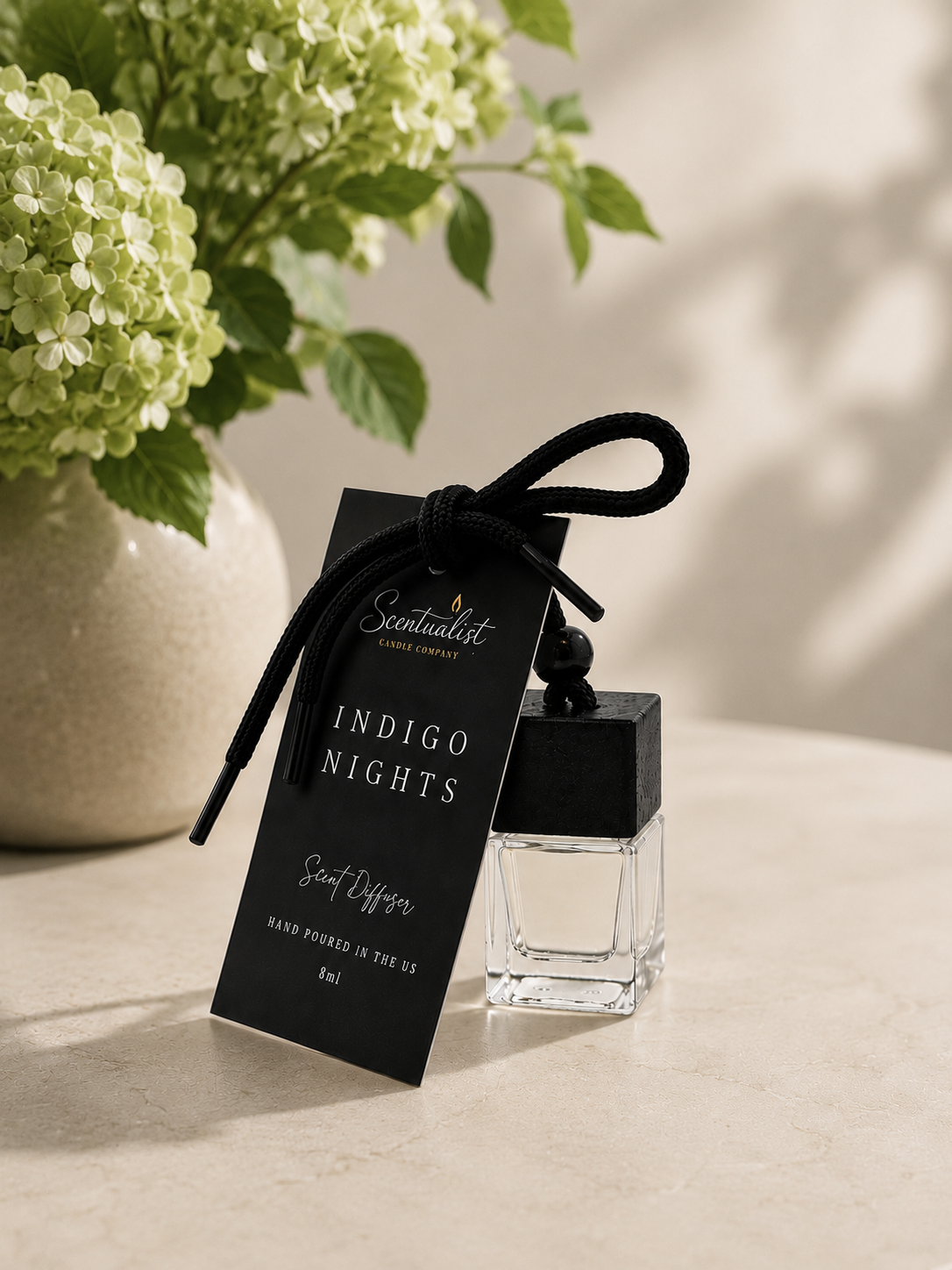 A scented candle diffuser labeled 'Indigo Nights' from Scentualist Candle Company, placed on a table next to a vase of green and white hydrangea flowers.