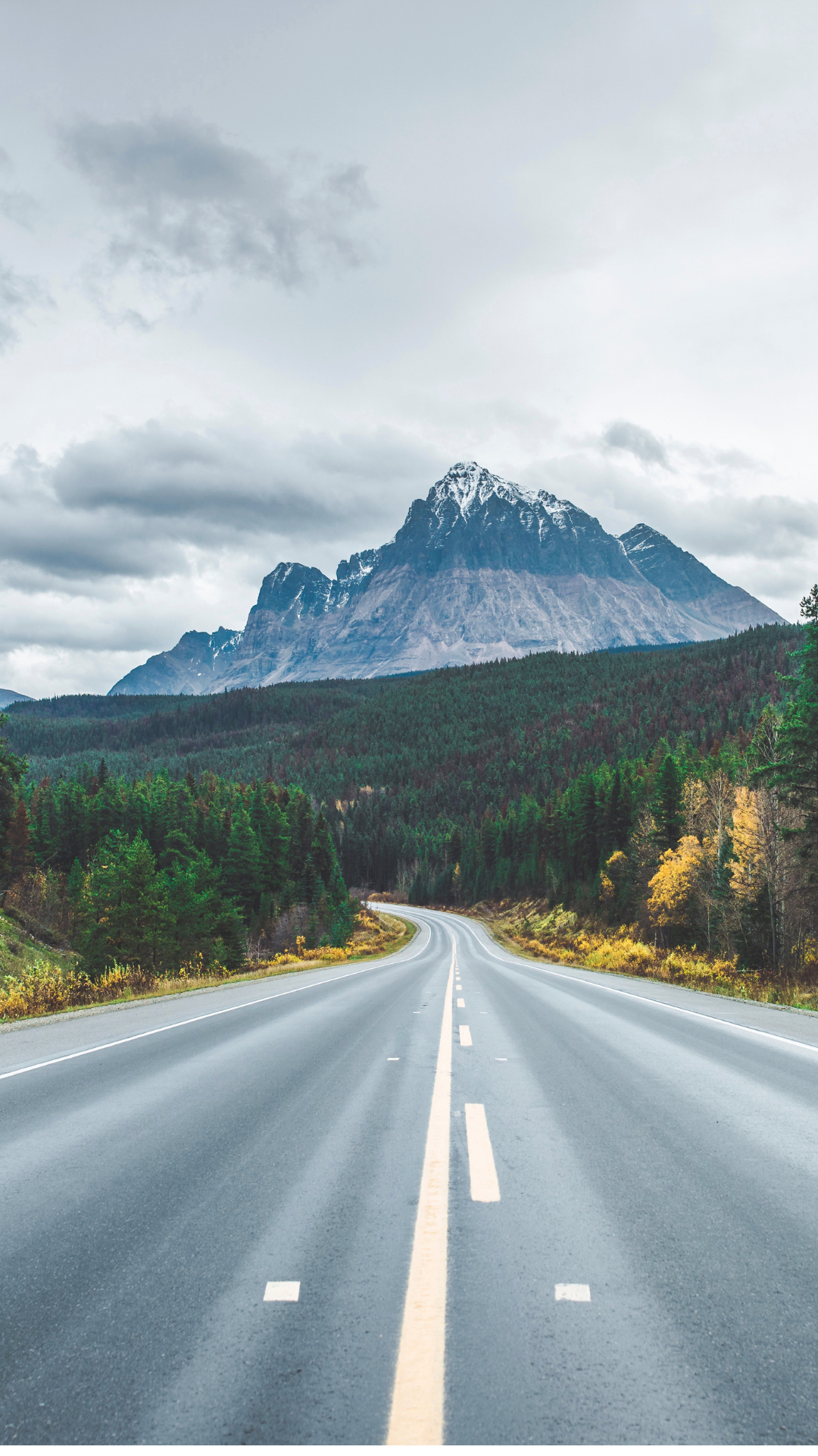 Empty two-lane road curving through a forested landscape with a mountain in the background and cloudy sky above.